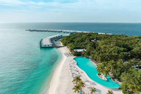 Luchtbeeld van een resort met een strand, een zwembad en bungalows op palen.