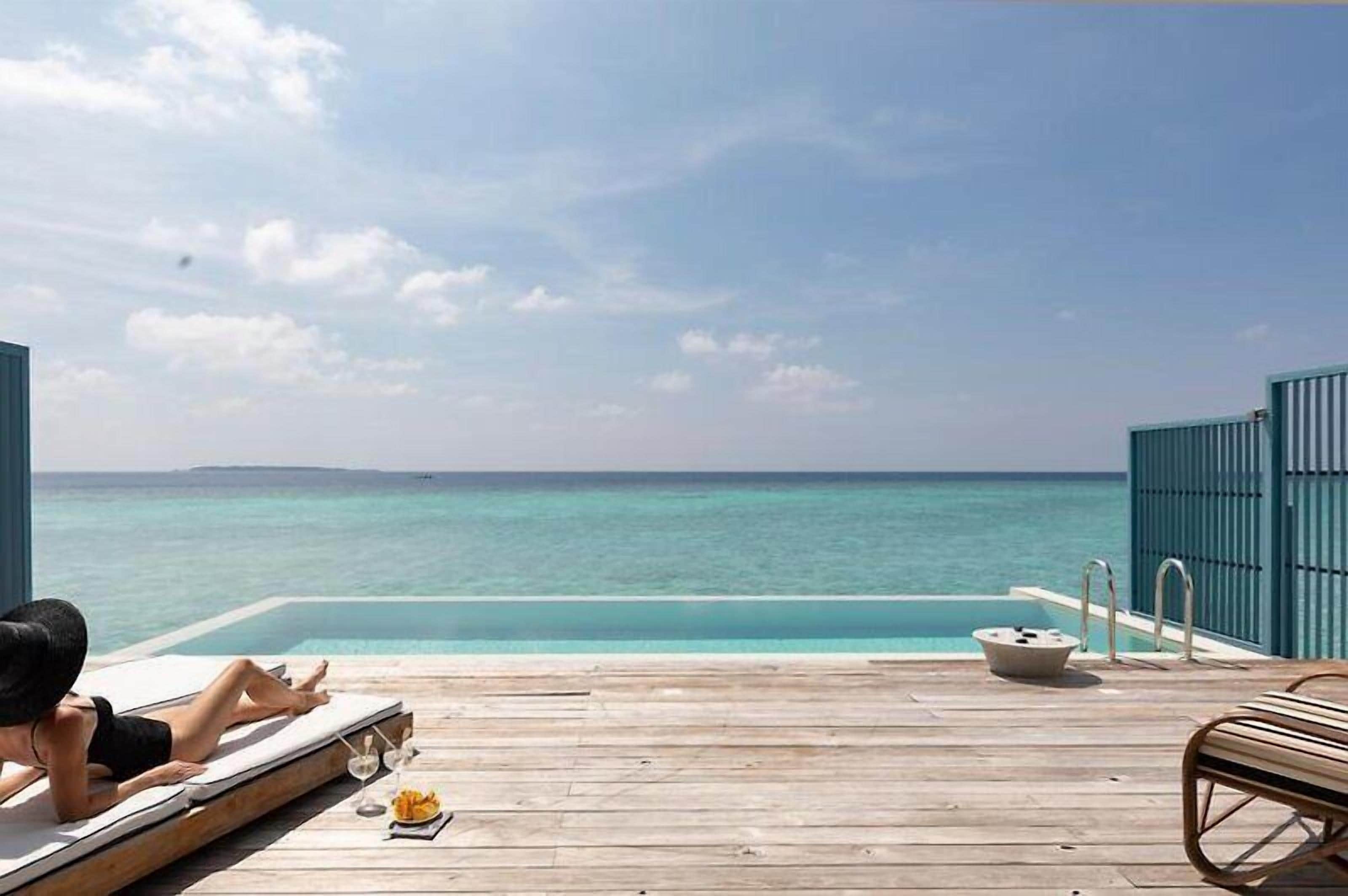 Una terraza de madera con piscina y sillones de descanso con vista a un mar azul y despejado.