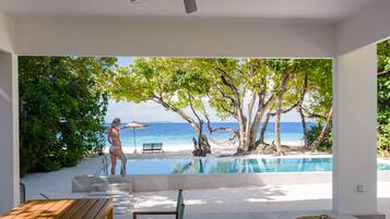 Quarto moderno com grande janela com vista para uma praia e uma piscina infinita.