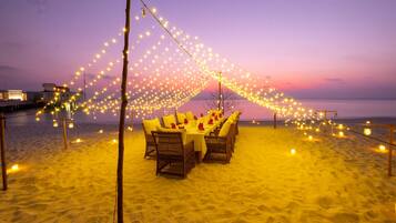 A beachside dining setup with tables and chairs, string lights, and a sunset in the background.
