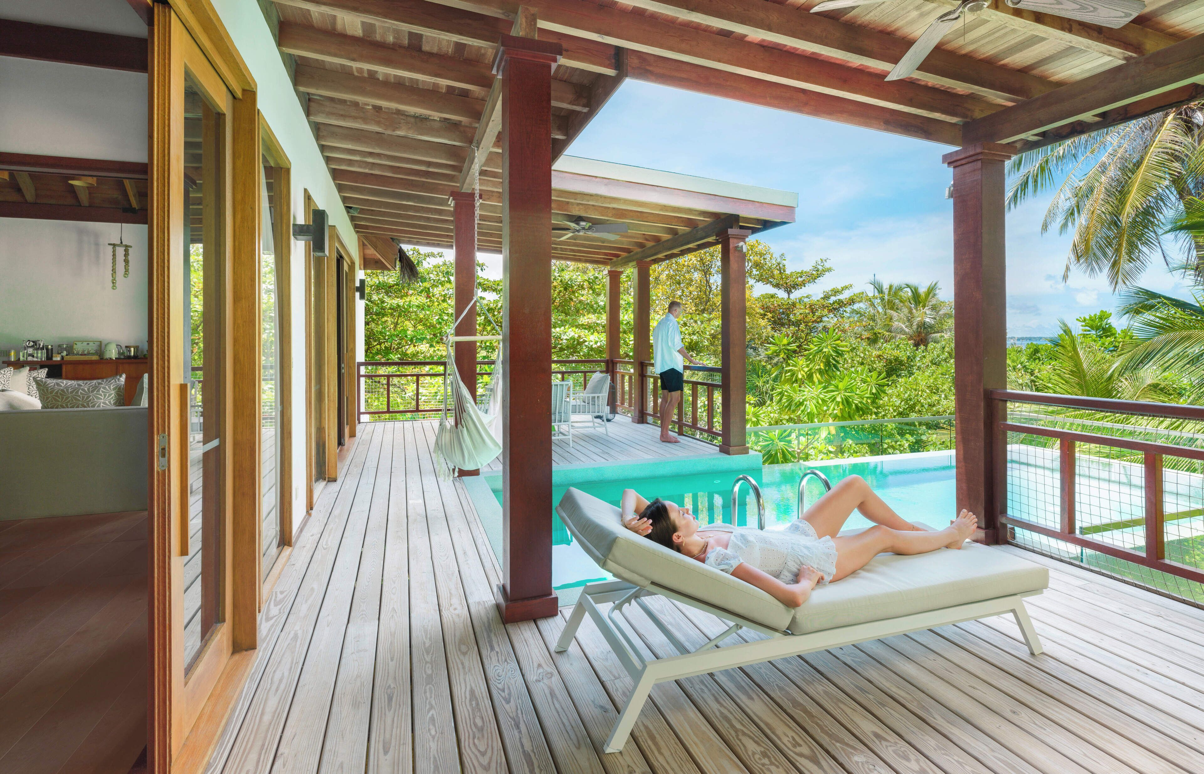 A wooden deck with a pool, lounge chairs, and a view of the ocean and greenery.