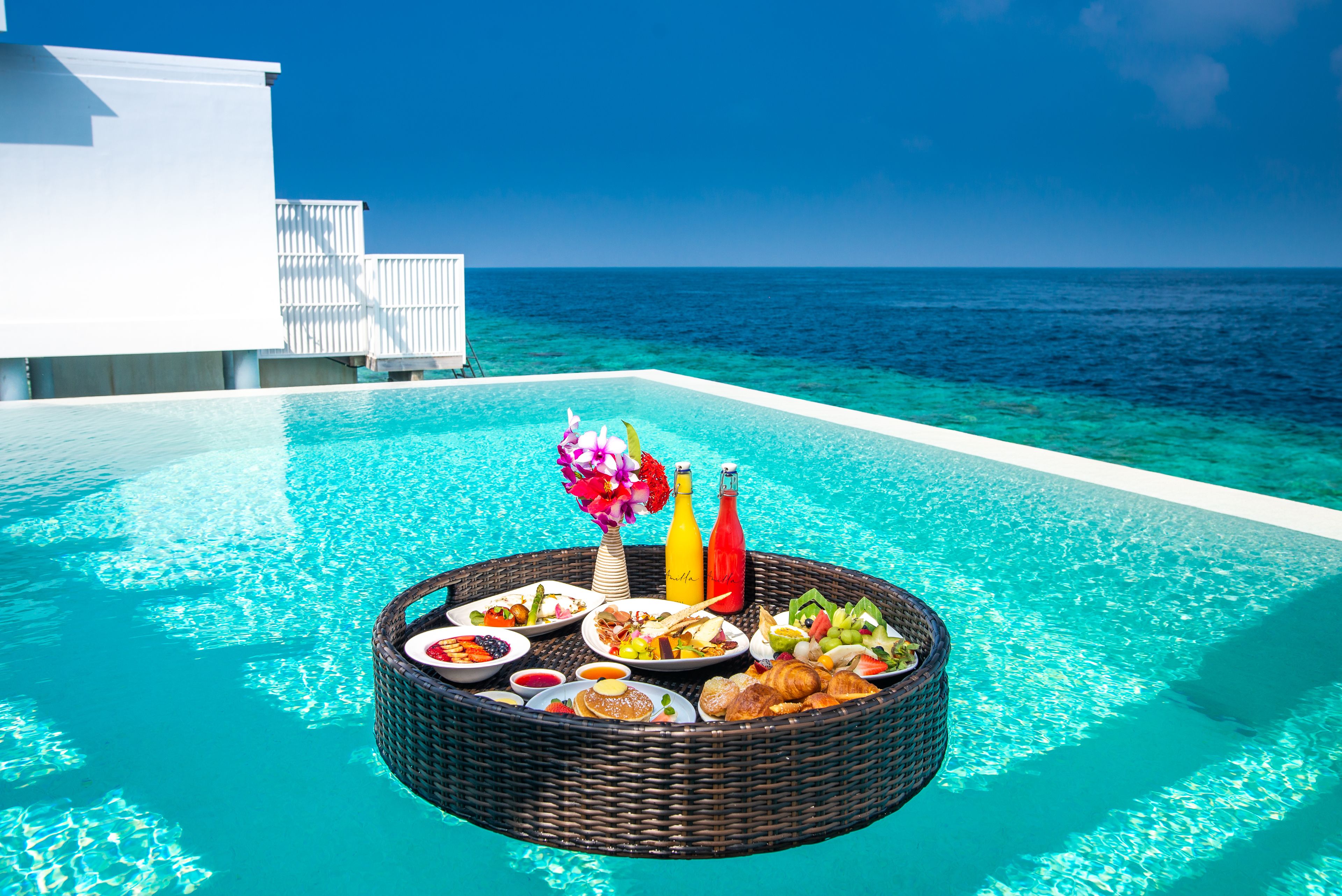 Desayuno junto a la piscina, con una mesa de mimbre, botellas y un arreglo floral.