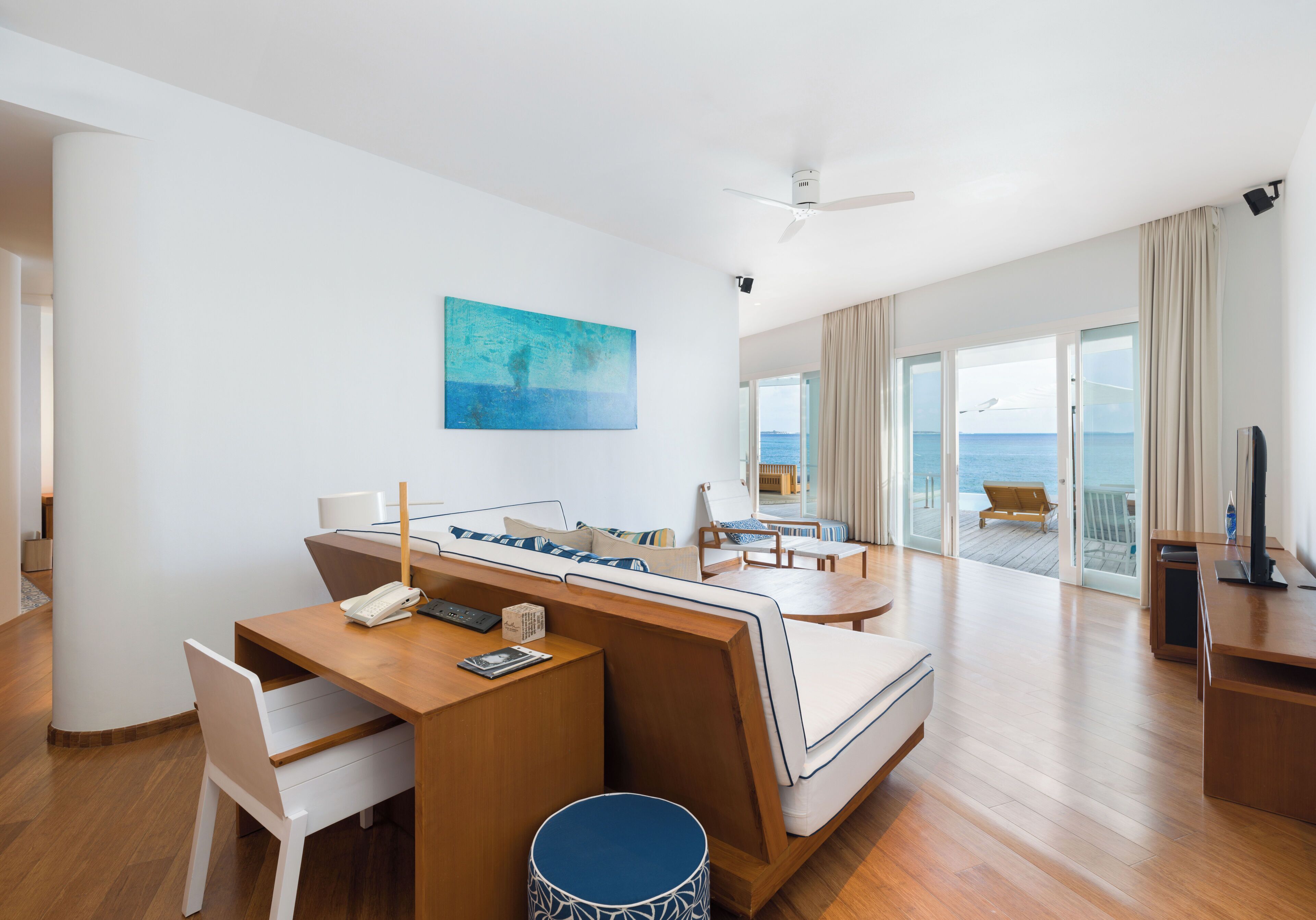 A modern hotel room with a wooden desk, a sofa, a television, and a large window offering a view of the sea.
