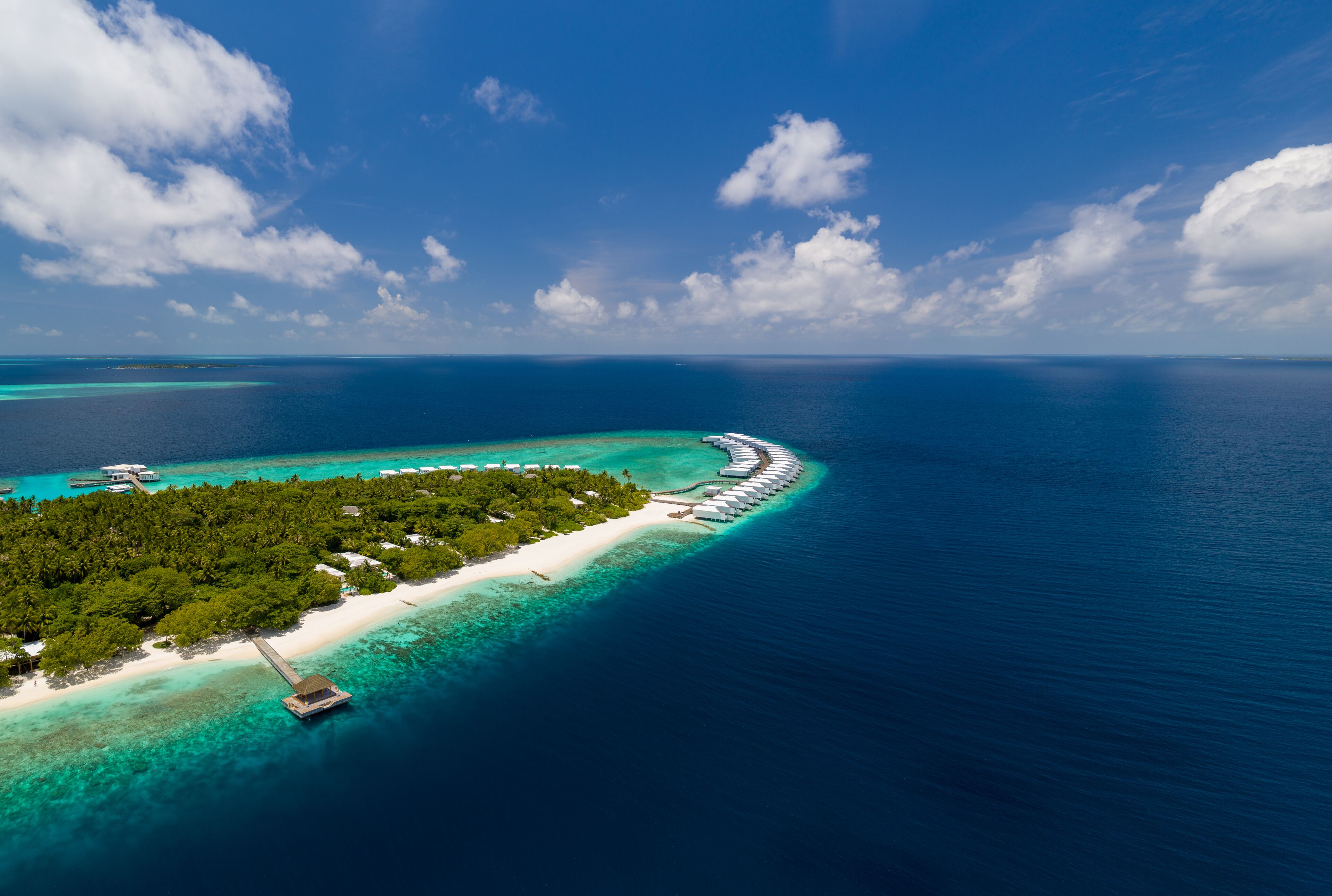 An aerial view of a tropical island with a long stretch of overwater bungalows, clear turquoise waters, and lush greenery.