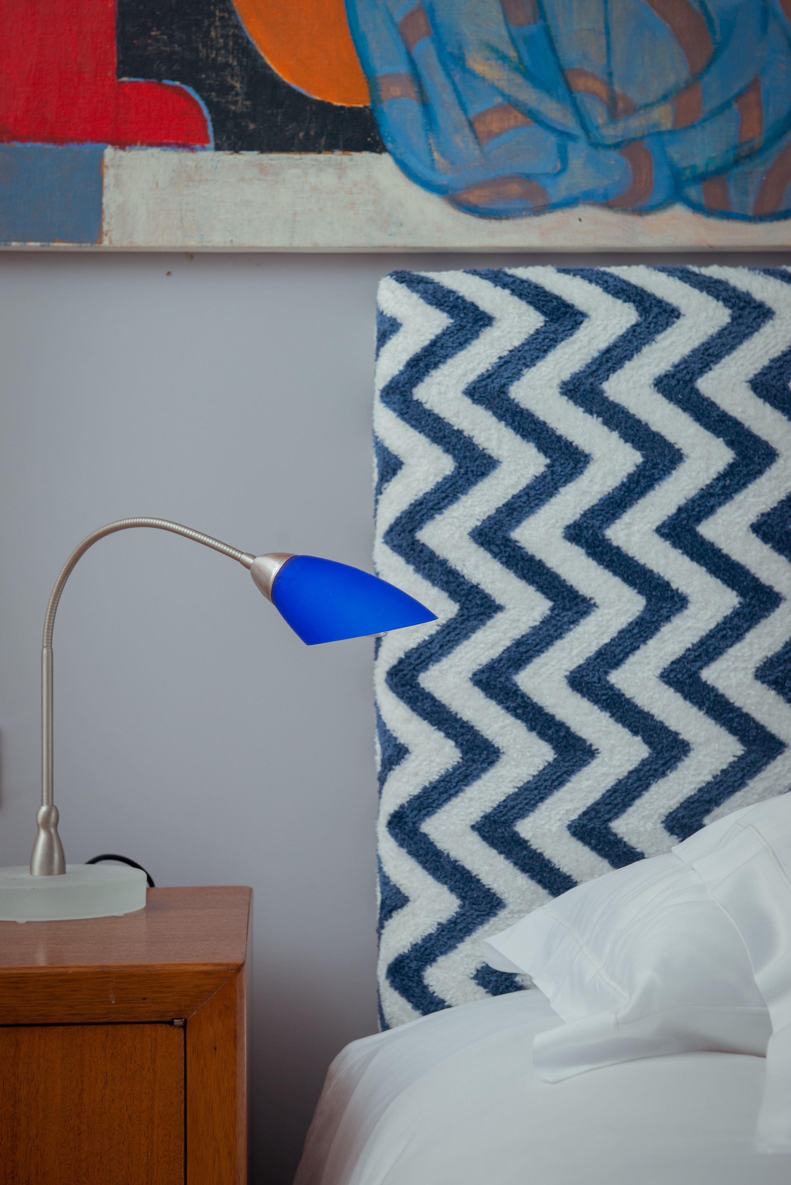 Una cama con almohada blanca y cabecera de tablero azul y blanco con diseño de chevrón. A la izquierda de la cama, una mesita de noche de madera con una lámpara de escritorio azul.