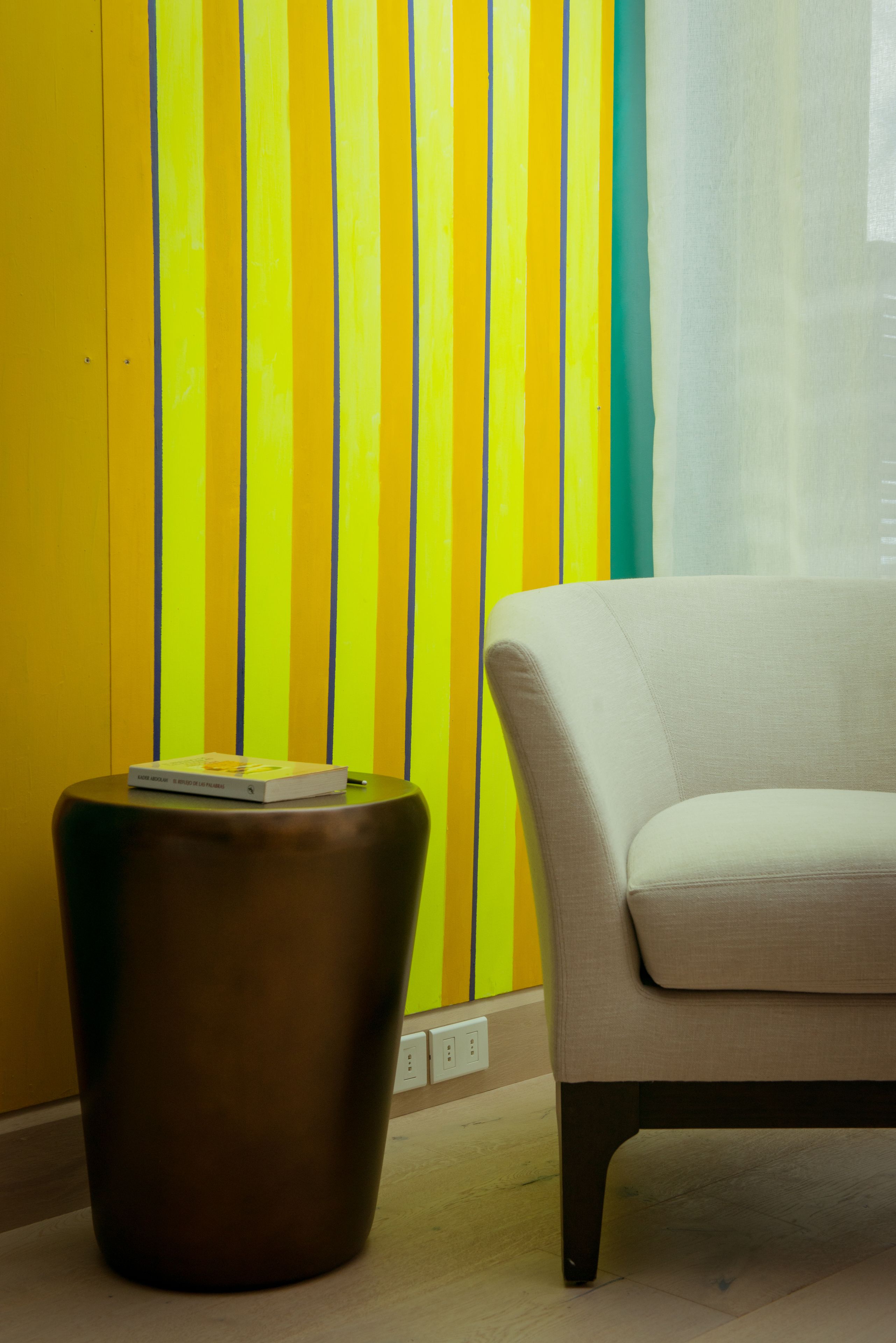 Una habitación con un sofá color beige, una mesita auxiliar marrón y una pared con rayas verticales en amarillo, verde y blanco.