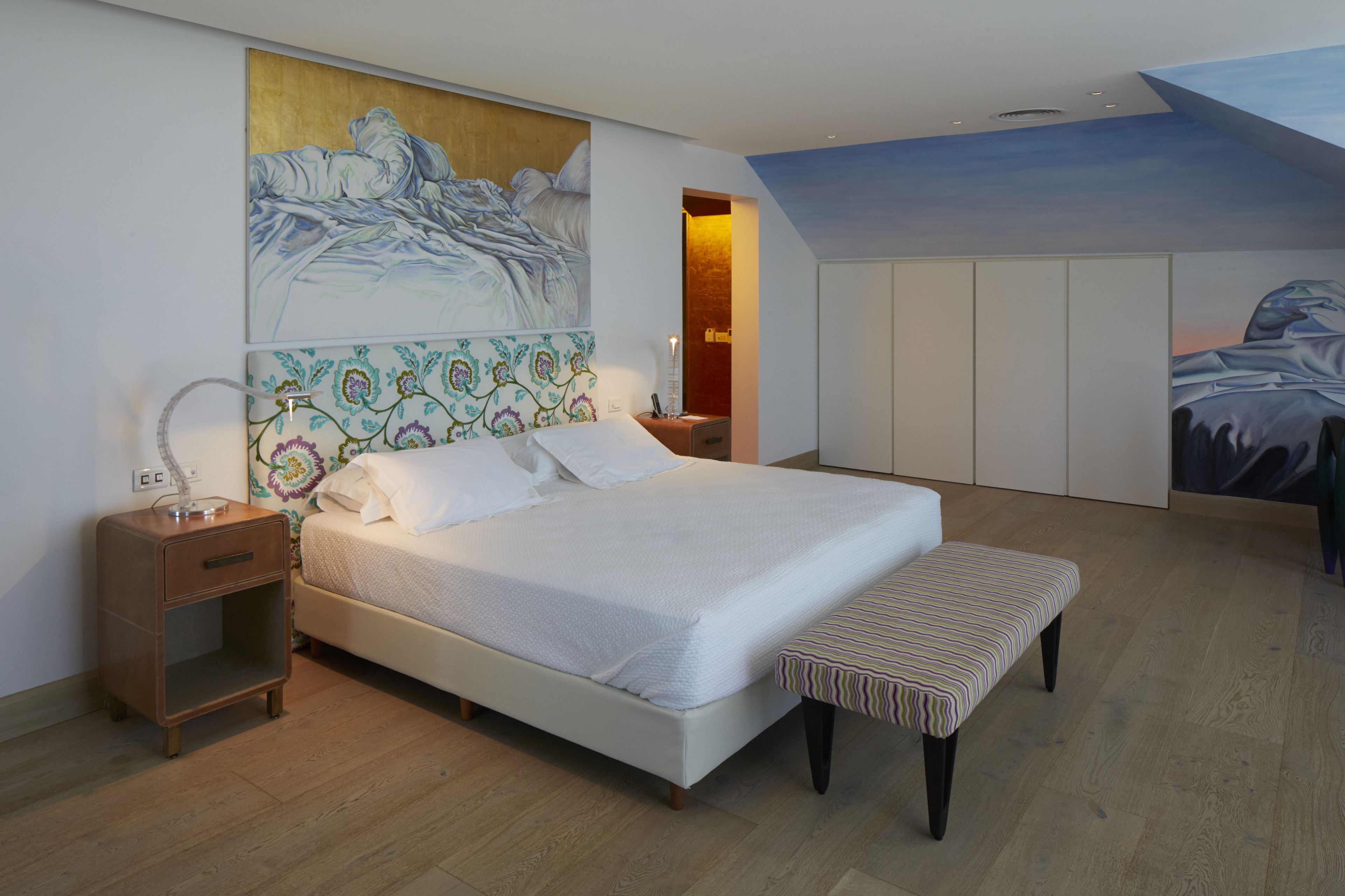Une chambre à coucher avec un grand lit, une table de chevet, un banc, une armoire et une fresque murale.