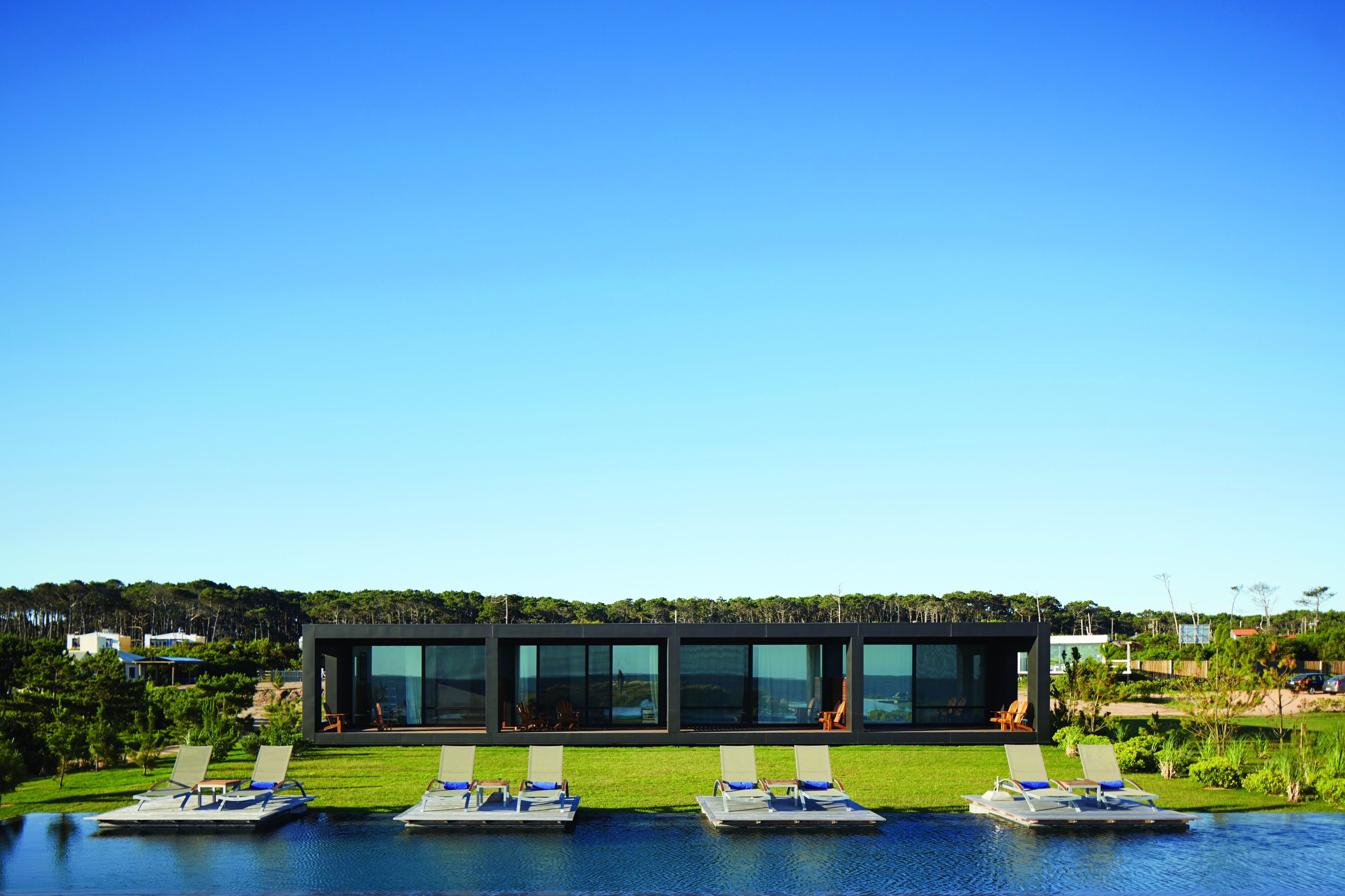 A modern building with a glass facade, a pool area with lounge chairs, and a view of a forested hill.