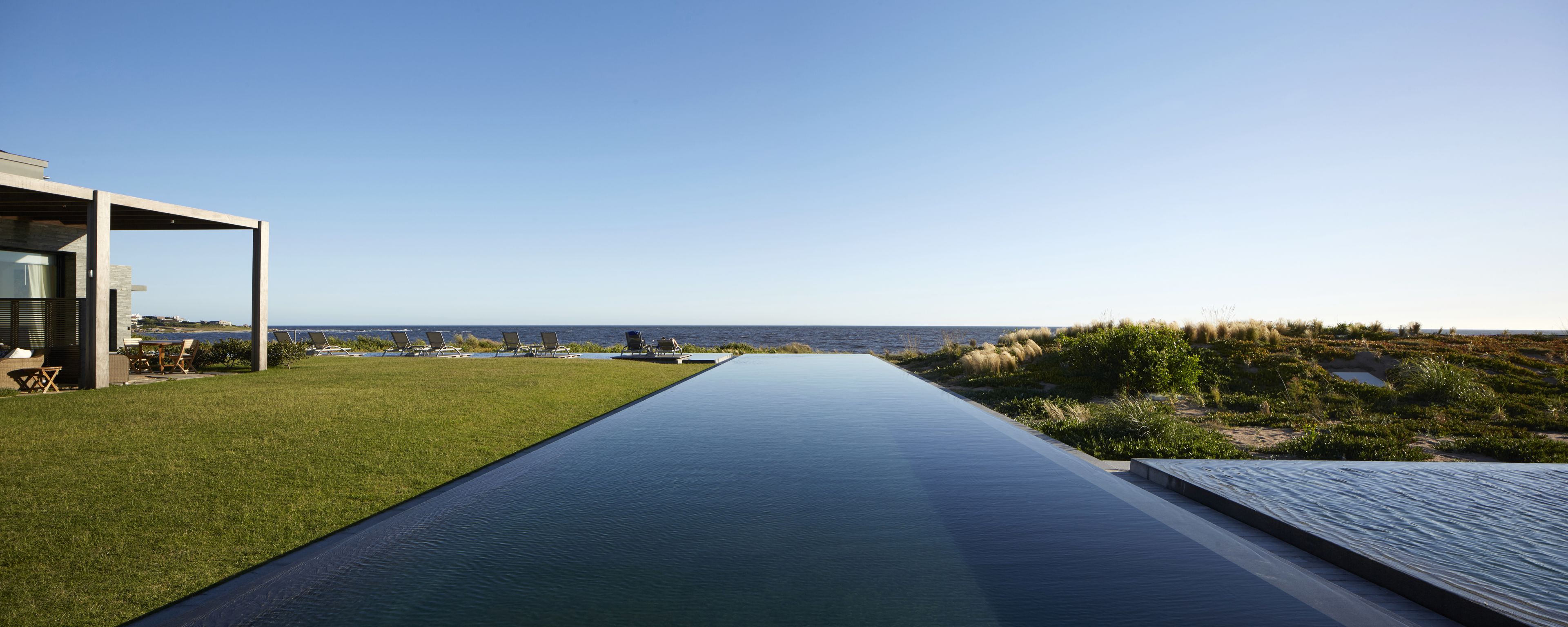 A modern infinity pool with a clear view of the ocean, a grassy area with lounge chairs, and a minimalist building structure.