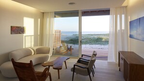 A modern living room with a curved sofa, wooden chairs, a coffee table, and a view of the beach through large glass doors. - Bahia Vik Jose Ignacio (José Ignacio)