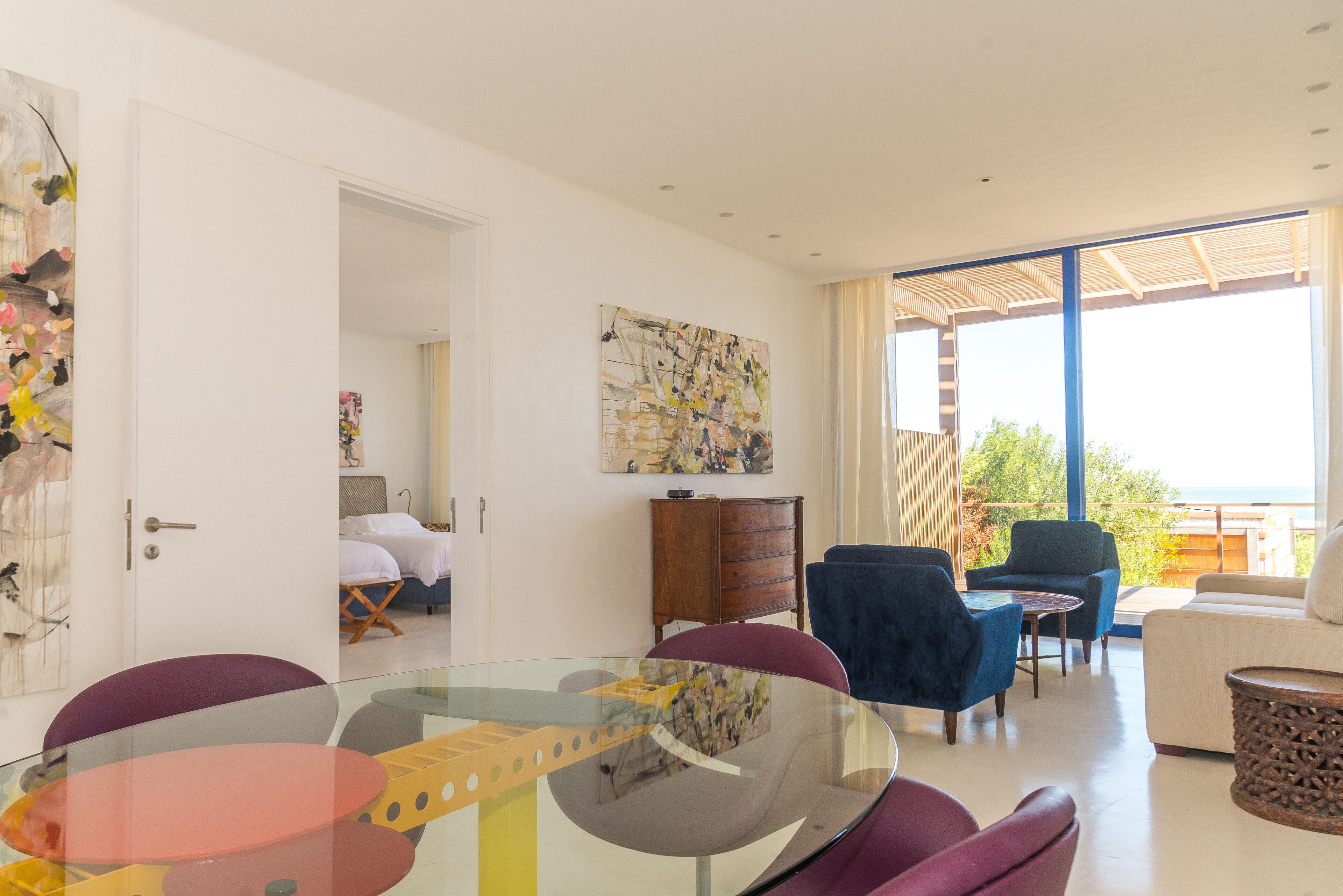 A modern living room with a glass dining table, purple chairs, a wooden cabinet, and a large window with a view of the sea.