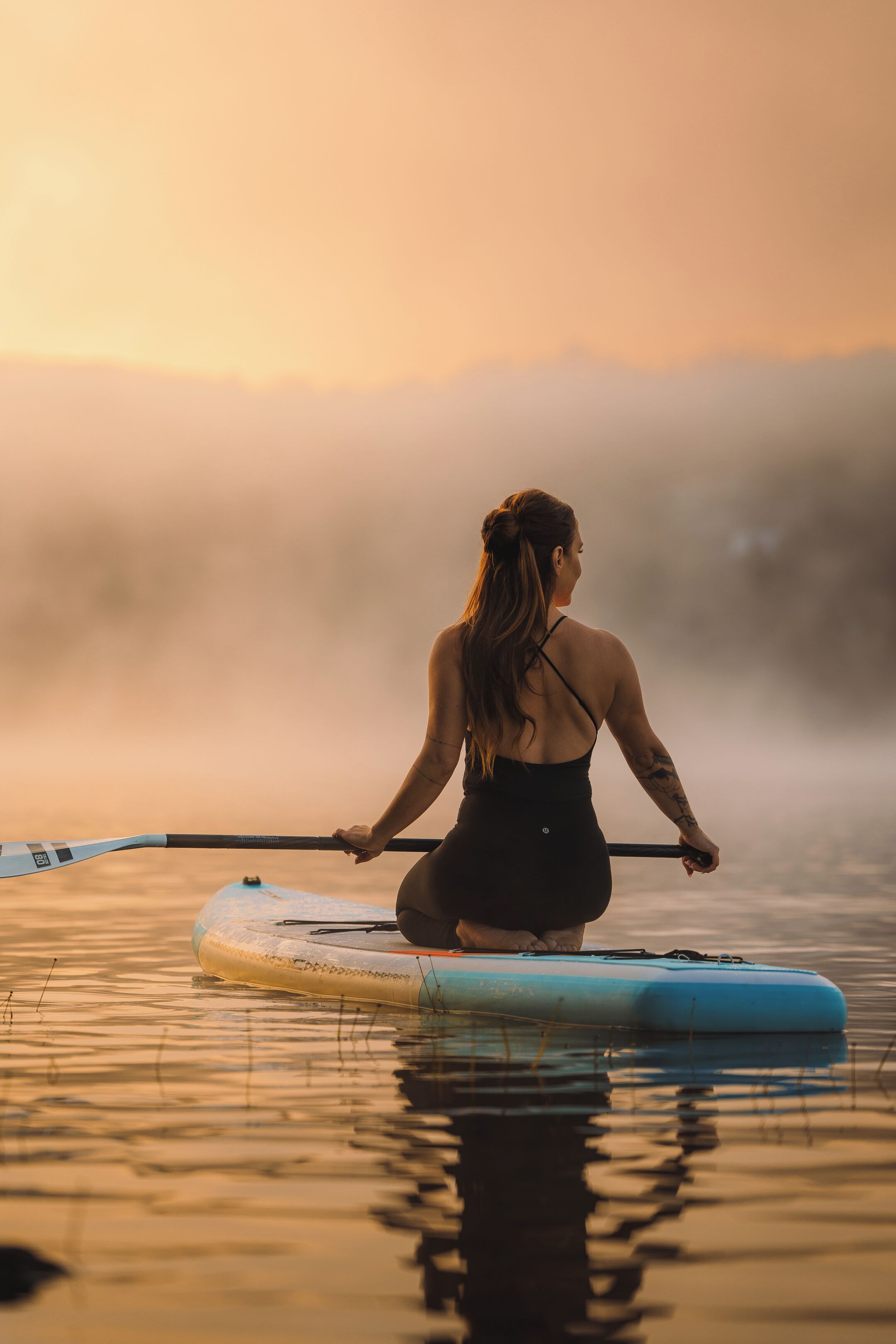 A person sitting on a paddleboard in calm water with a misty background.