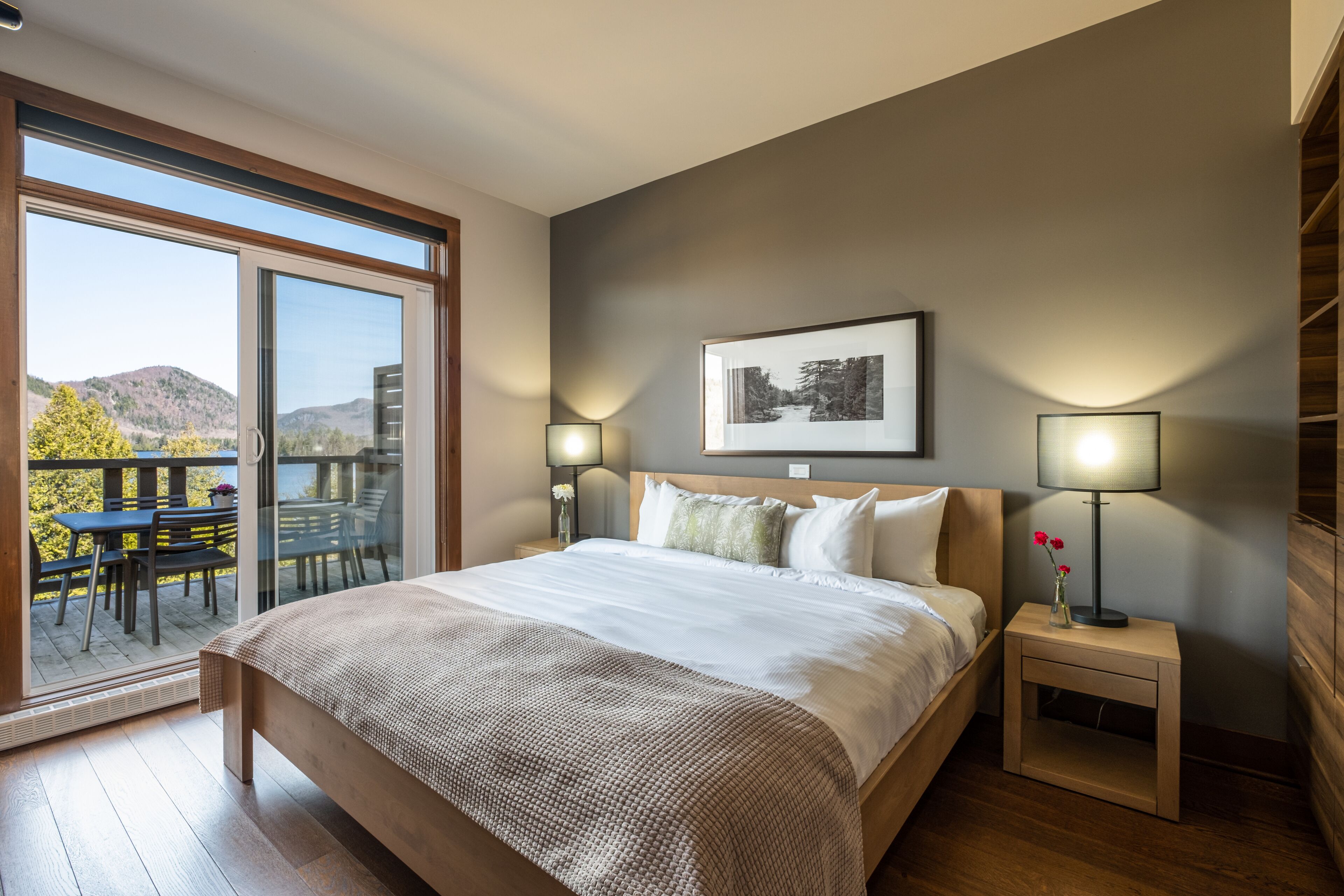 Un dormitorio con una cama grande, una mesita de noche con una lámpara y vistas a una terraza y montañas.