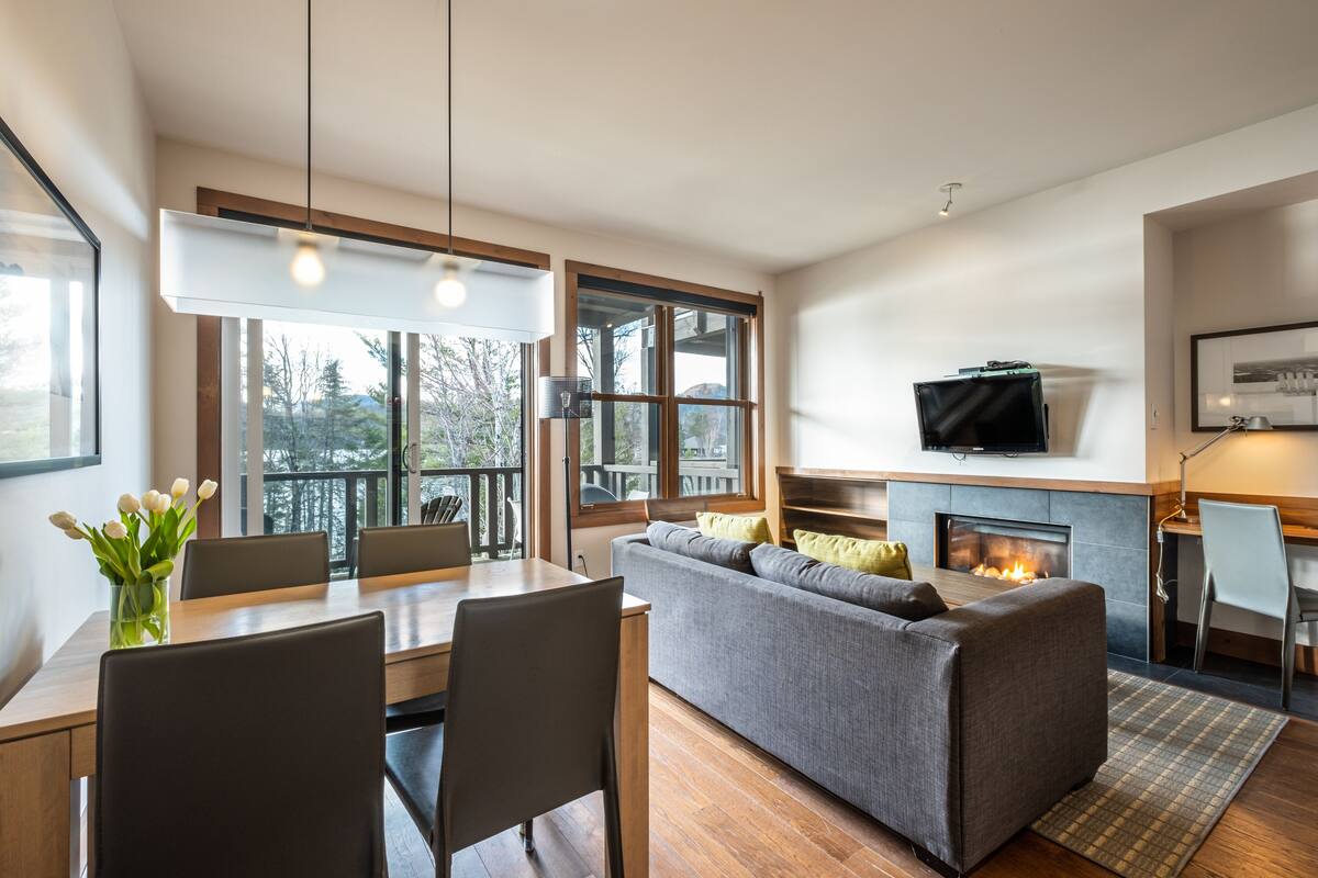 Un salon moderne doté d’une cheminée, d’un canapé, d’une table à manger avec des chaises et de grandes fenêtres offrant une vue sur l’extérieur.