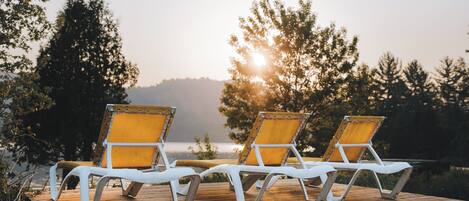 Deux fauteuils de jardin blancs avec des coussins jaunes, placés sur une terrasse en bois donnant sur une étendue d’eau, avec des arbres en arrière-plan.