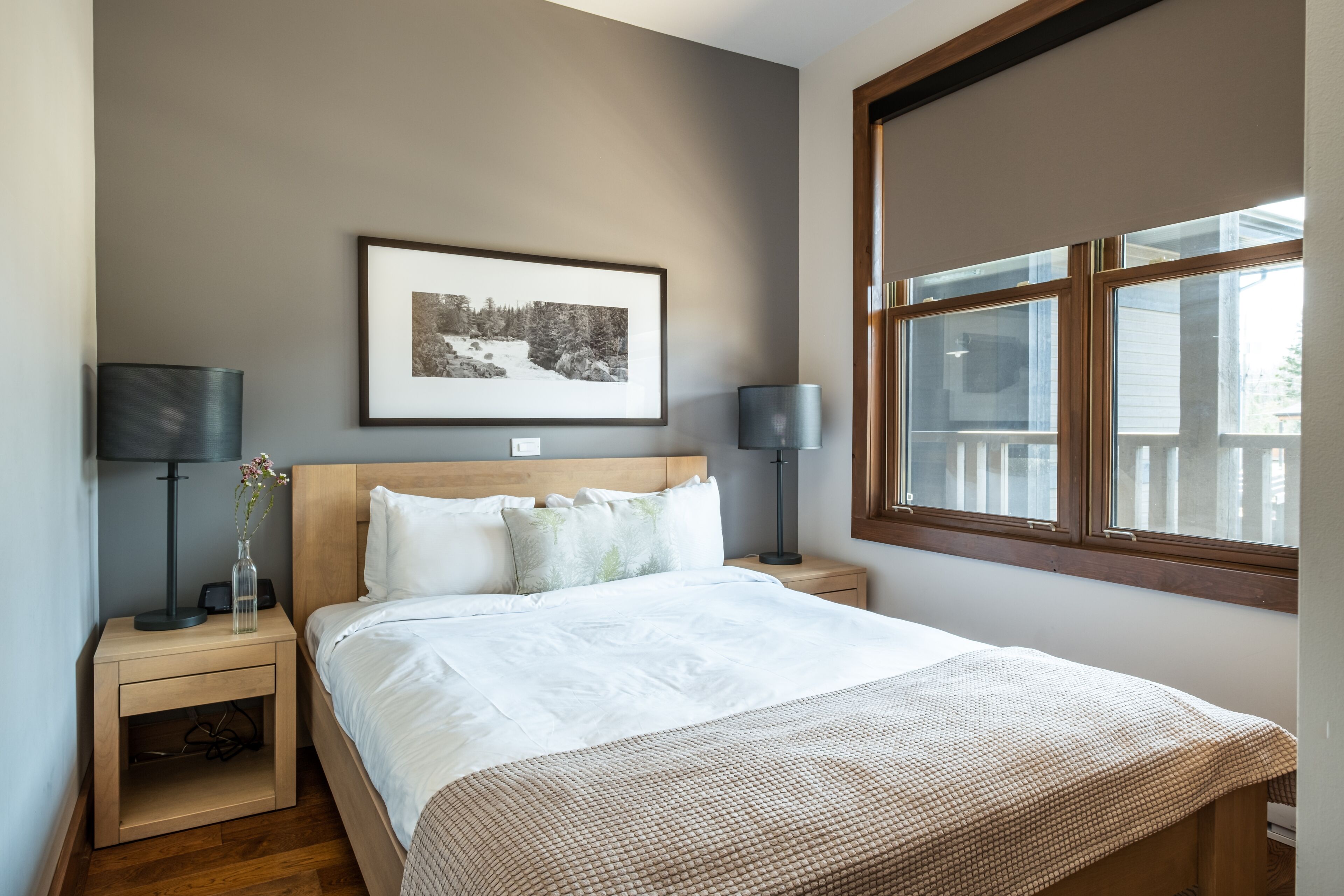 A neatly arranged bedroom with a large bed, two bedside tables with lamps, a framed picture on the wall, and a window providing natural light.