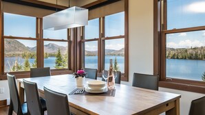 A dining area with a table set for six, large windows with a lake and mountains view, and a modern pendant light. - Suites Sur Lac (Lac-Superieur)