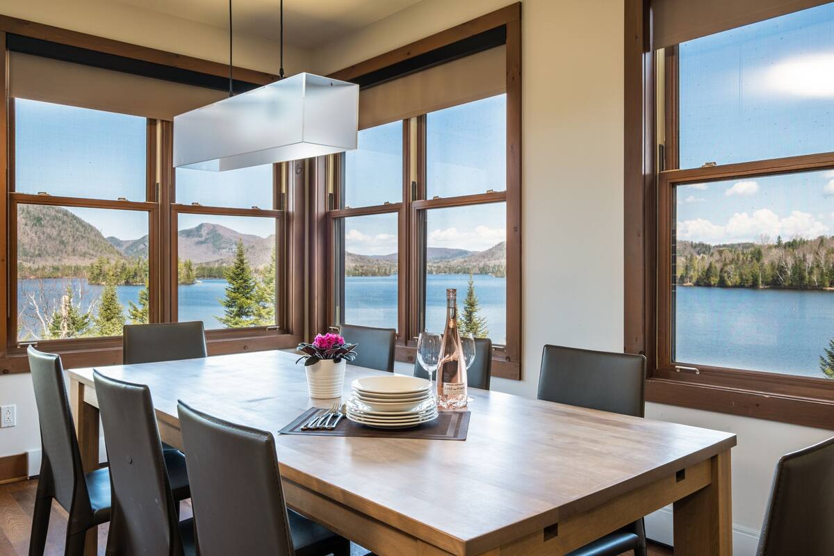 Un coin repas avec une table dressée pour six personnes, de grandes fenêtres offrant une vue sur le lac et les montagnes, et un luminaire suspendu moderne.