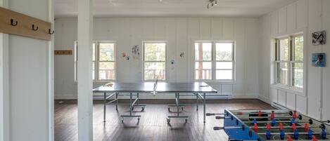 Una habitación amplia con mesa de futbolín, mesa de ping pong y ventanas grandes que dejan entrar luz natural.