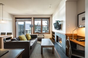 A modern living room with a sofa, coffee table, and fireplace. There are large windows and a television mounted on the wall. - Suites Sur Lac (Lac-Superieur)