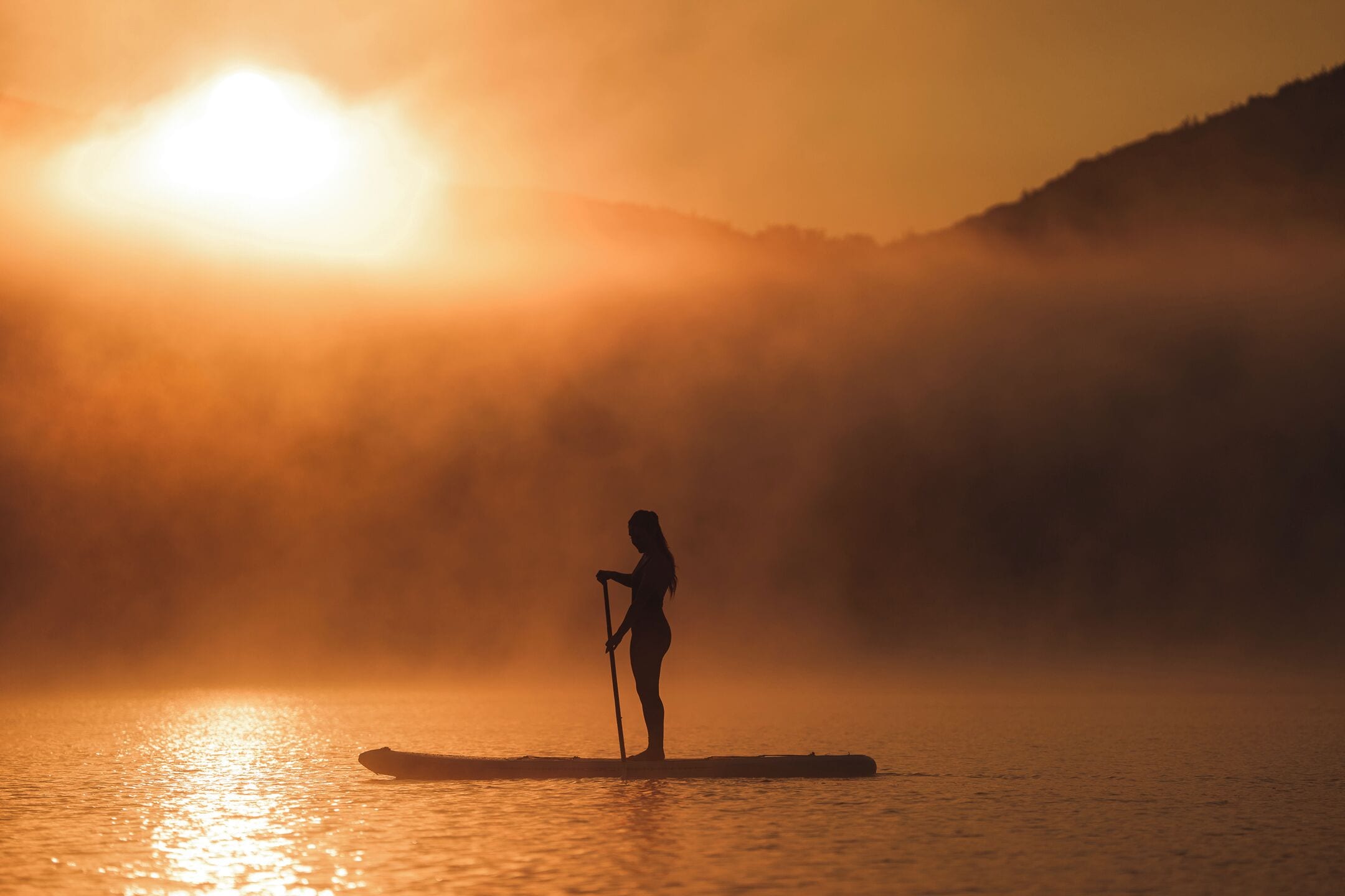 Una persona fa paddleboarding su un'acqua calma all'alba, con montagne nebbiose sullo sfondo.