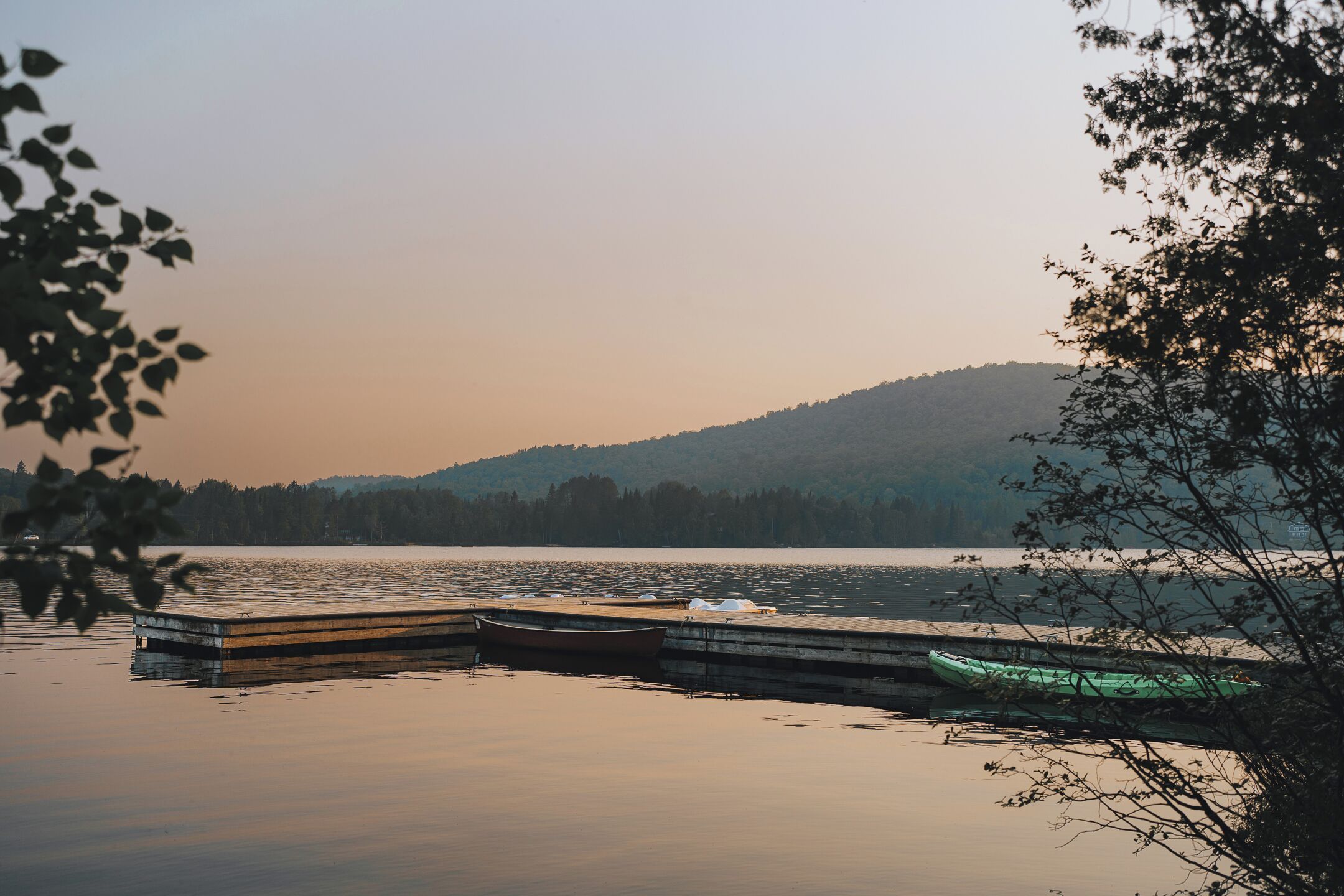 A calm lake with a dock and boats, surrounded by trees and hills in the background.