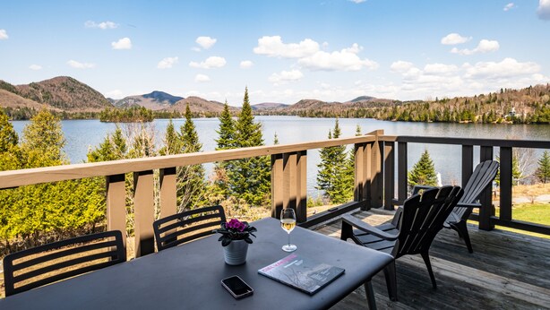A deck with a table, chairs, a potted plant, a glass of wine, and a magazine. A lake and mountains are visible in the background. - Suites Sur Lac (Lac-Superieur)