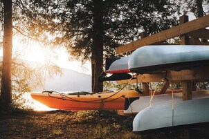 Kayak y canoa guardados bajo una estructura de madera, probablemente un cobertizo, con un paisaje natural de árboles y una montaña al fondo.