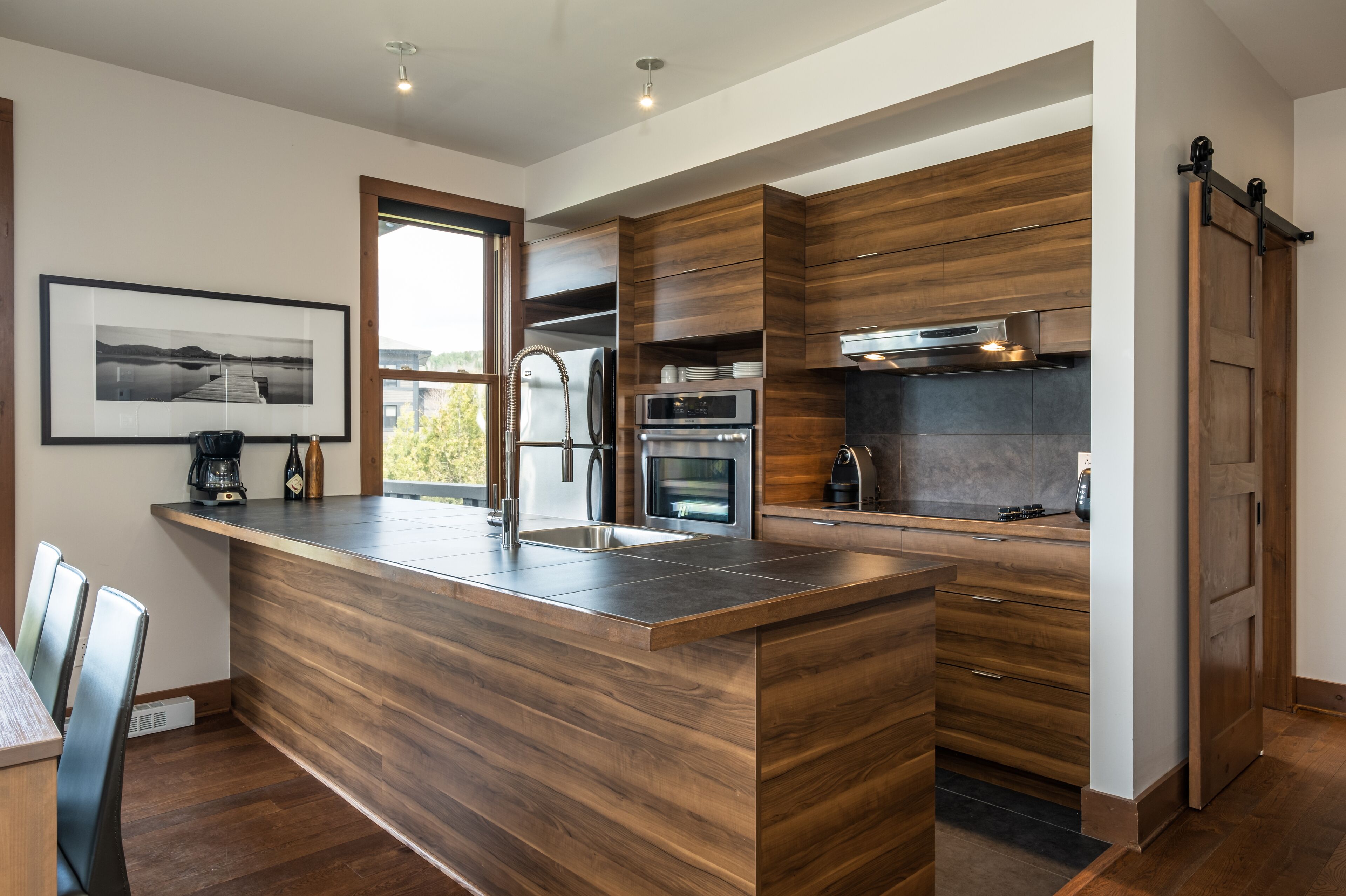 Una cocina moderna con armarios de madera, una isla central con lavamanos, electrodomésticos integrados y un amplio ventanal que proporciona luz natural.