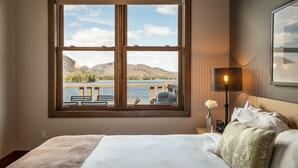 A bedroom with a large window overlooking a lake and mountains, a bed with white linens, a nightstand with a lamp, and a framed picture on the wall. - Suites Sur Lac (Lac-Superieur)