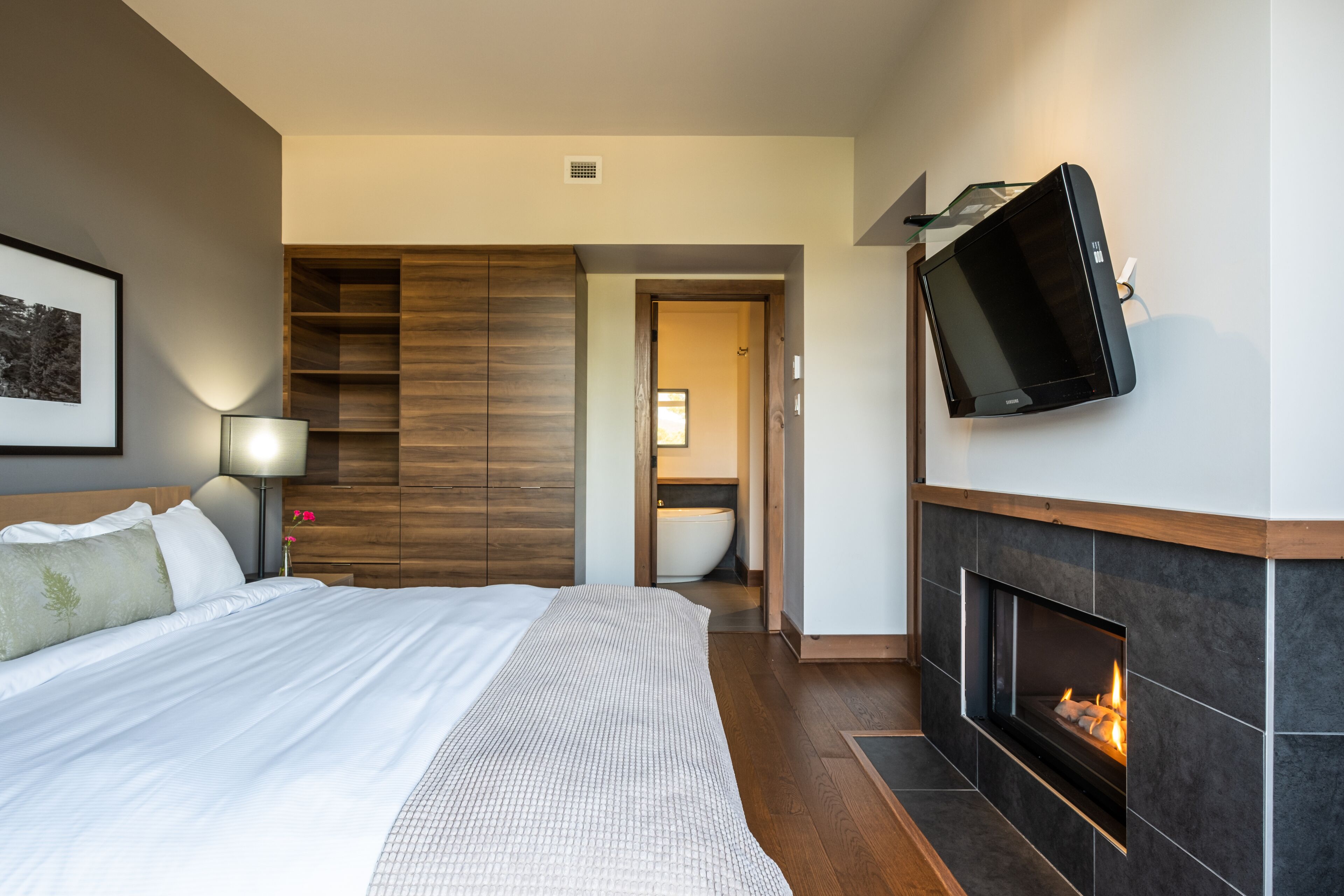 Una habitación de hotel moderna con una cama grande, un televisor de pantalla plana montado en la pared, una chimenea y un armario de madera.