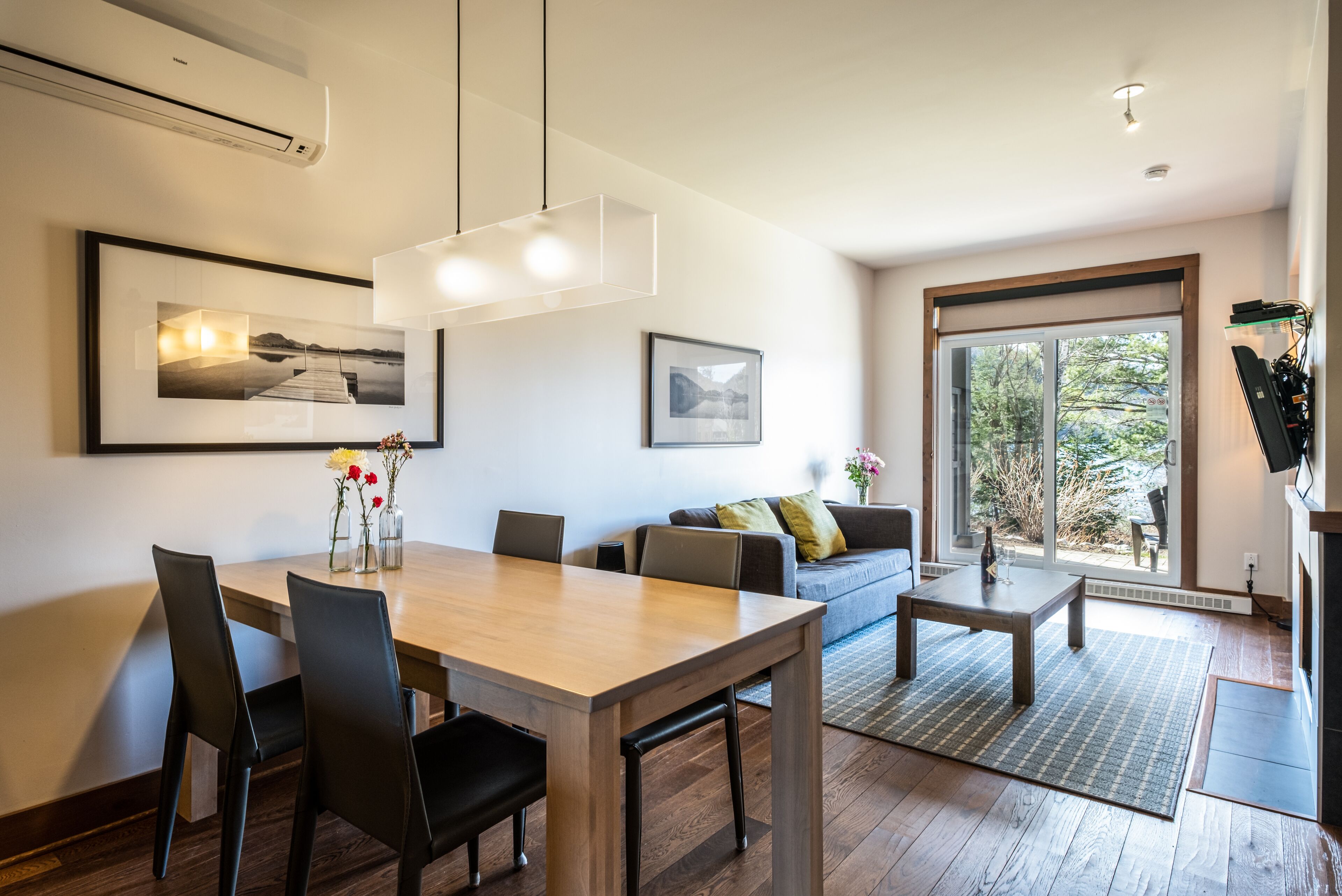 Una sala de estar moderna con una mesa de comedor de madera, sillas negras, un sofá y una mesa de centro. Hay cuadros en las paredes y un ventanal con vistas a árboles.