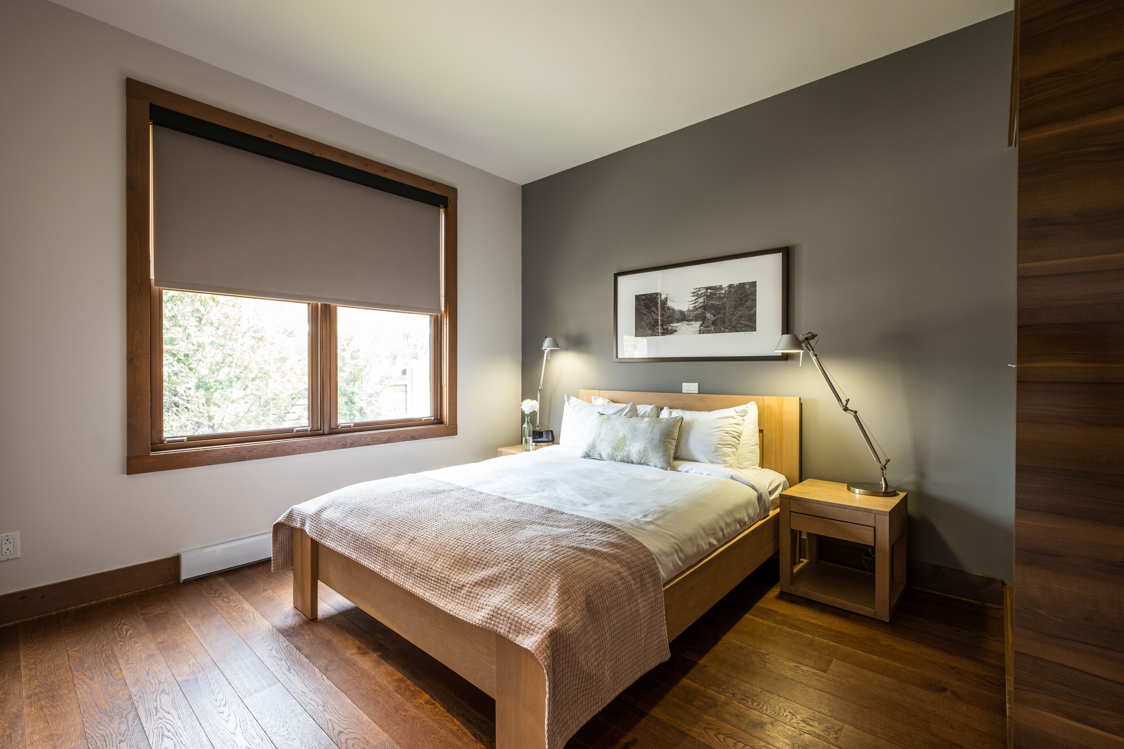 A bedroom with a wooden bed, a nightstand, a lamp, a framed picture on the wall, and a window with a view of trees.