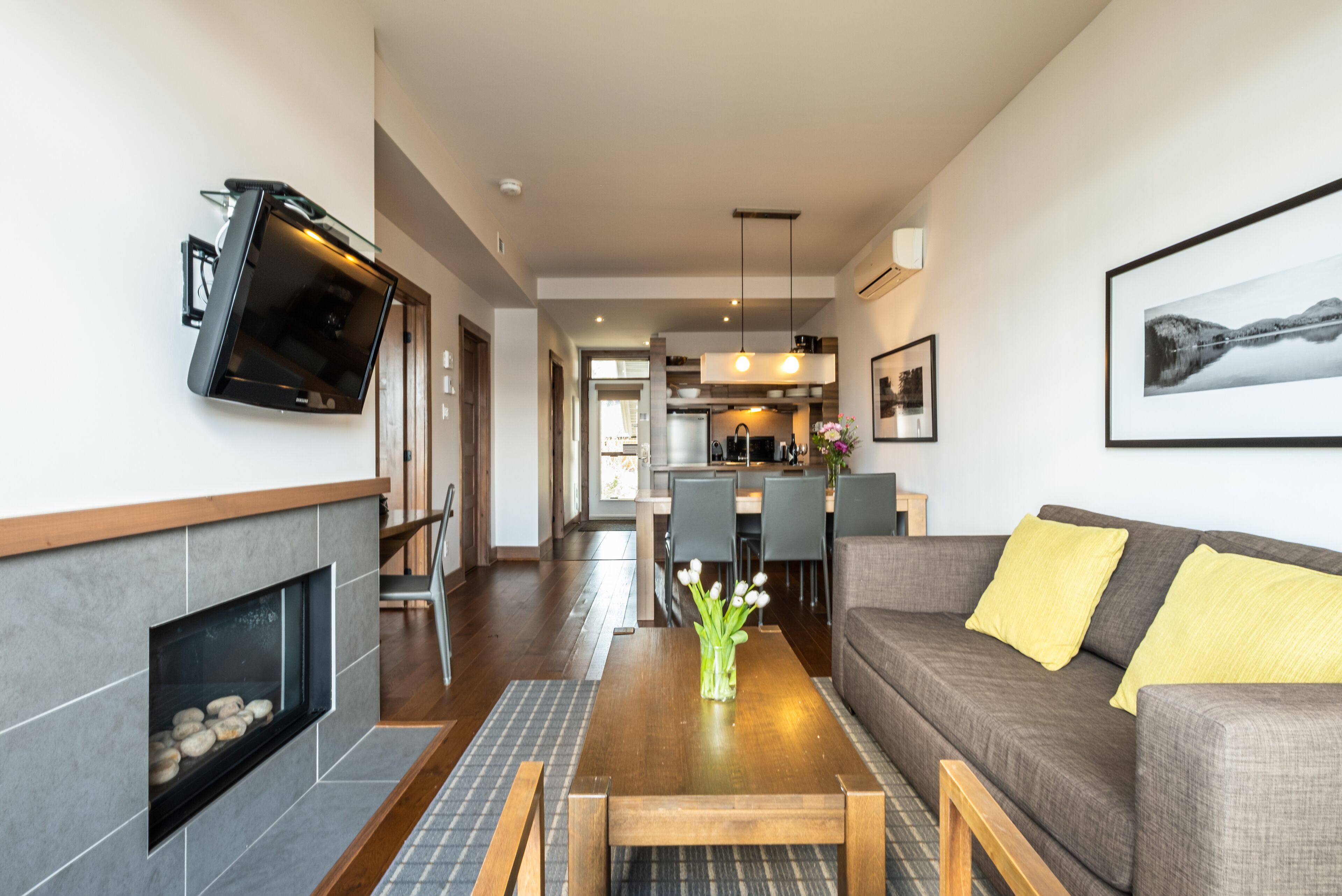 A modern living room with a fireplace, a flat-screen TV mounted on the wall, a sofa with yellow cushions, and a dining area with chairs and a table.