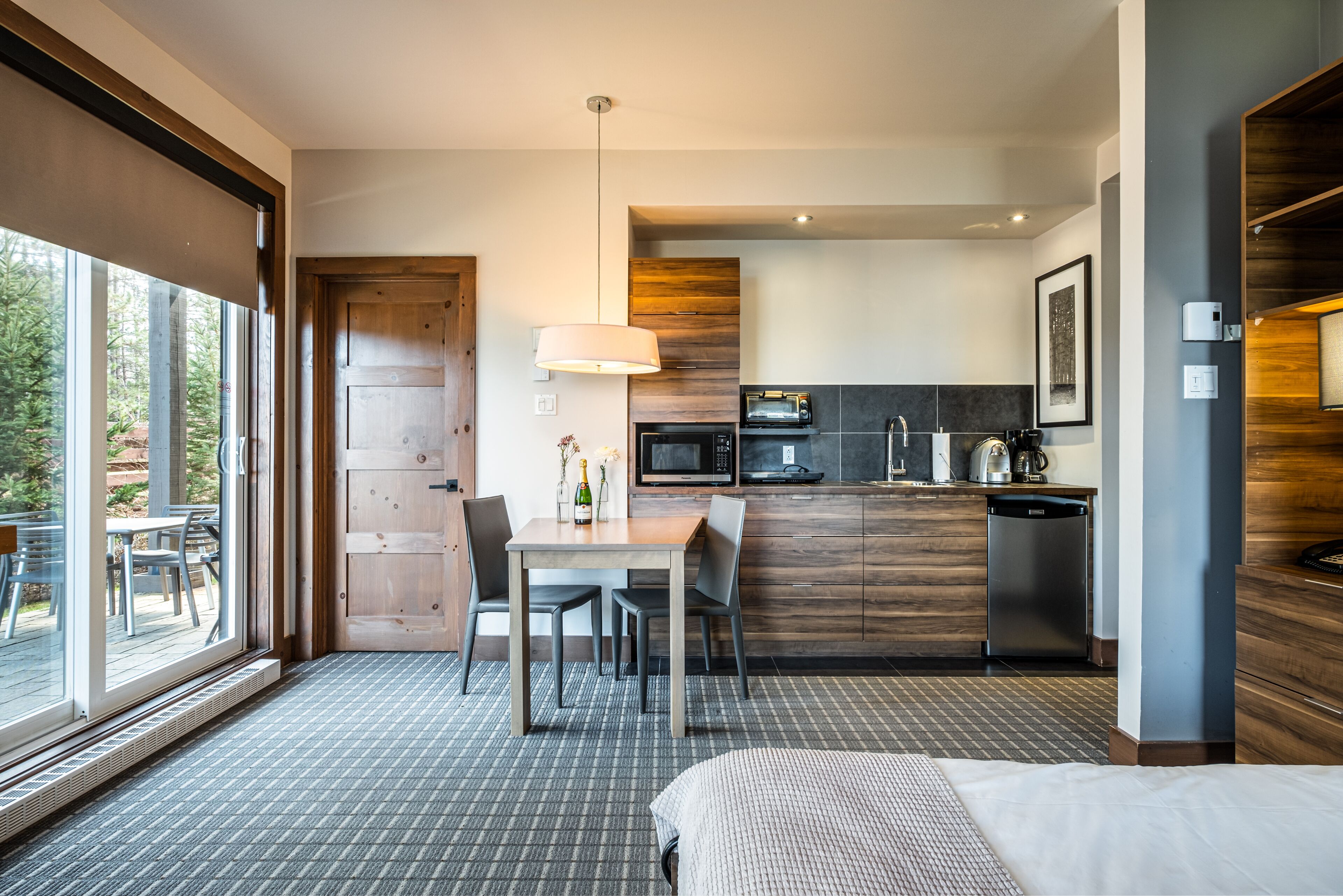 A modern hotel room with a large window, a dining area with a table and chairs, a kitchenette, and a bed.