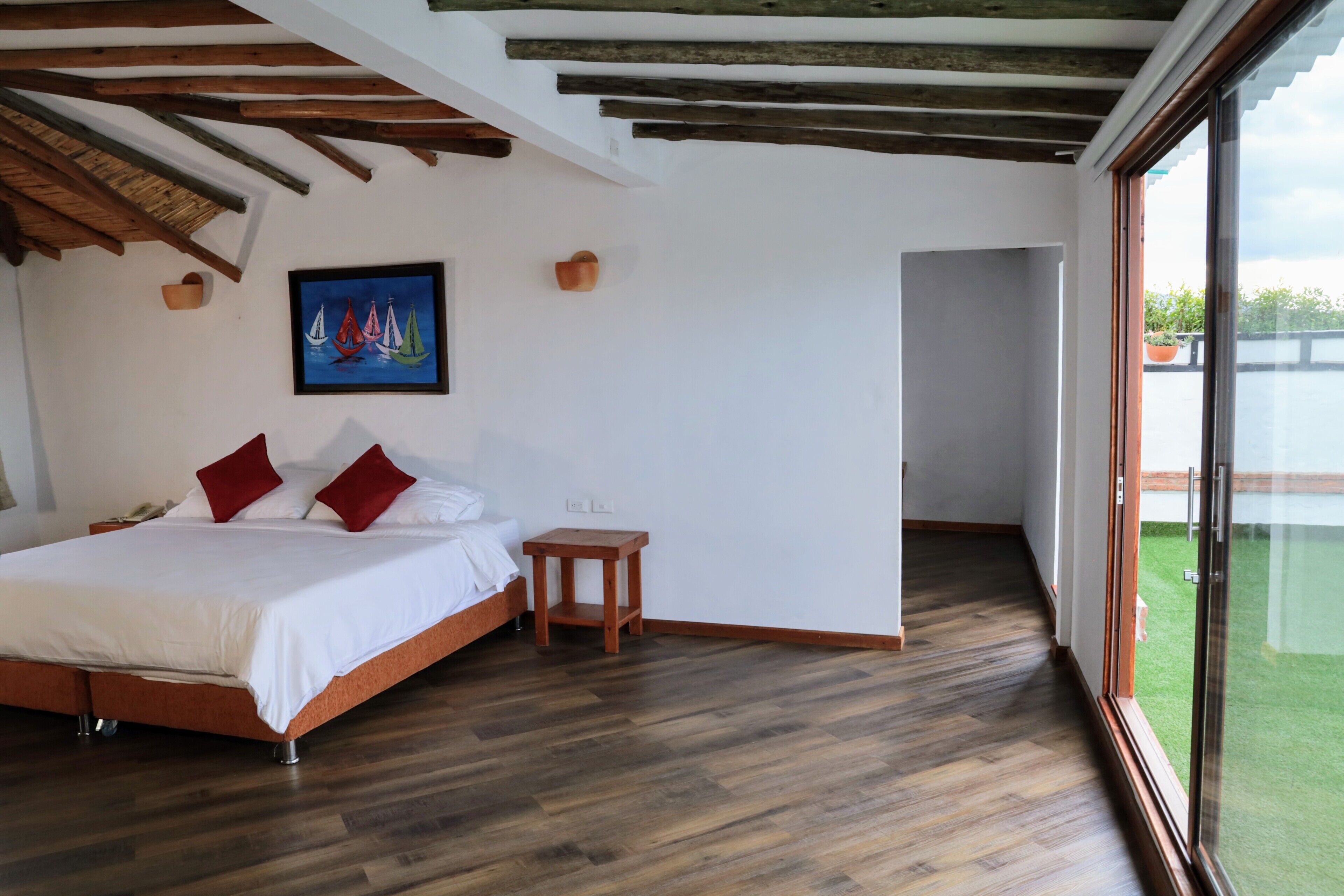 Une chambre avec un lit en bois, une petite table de chevet, un tableau représentant des voiliers et une vue sur une pelouse verdoyante, visible à travers une porte vitrée.