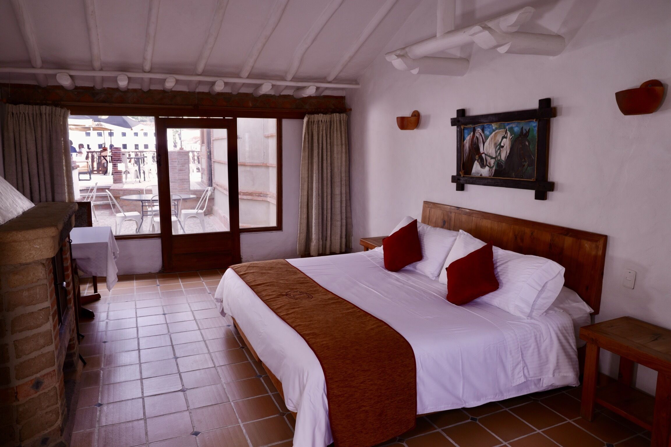 Habitación de hotel con una cama grande, cabecera de madera, un cuadro de caballos, una mesita y vistas al patio exterior.