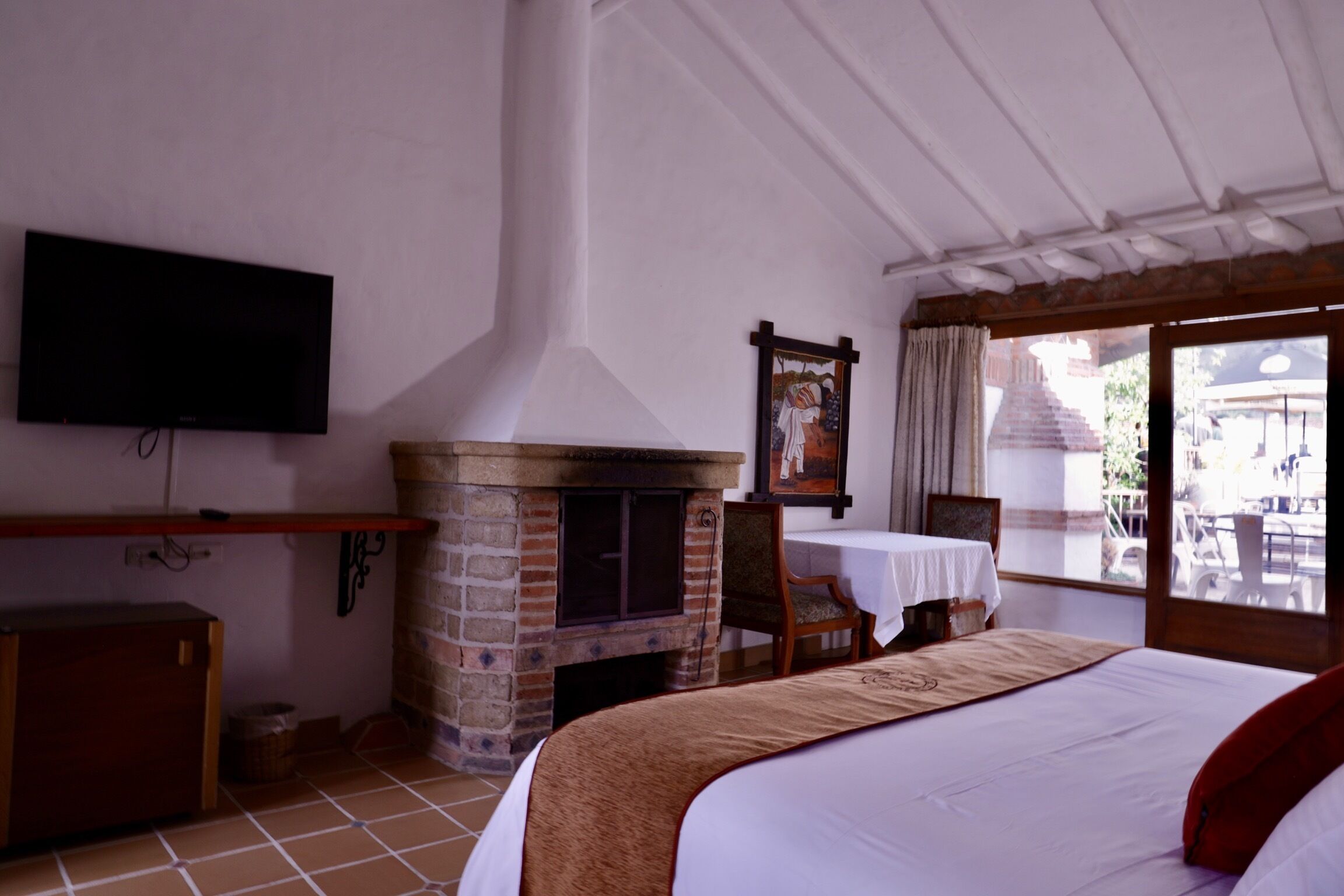 Ein Hotelzimmer mit Kamin aus Ziegelsteinen, einem an der Wand montierten Fernseher, einem Bett mit rotem Kissen und Blick durch große Fenster nach draußen.