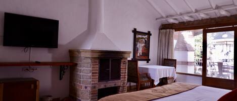 A hotel room with a brick fireplace, a television mounted on the wall, a bed with a red pillow, and a view of the outside through large windows.