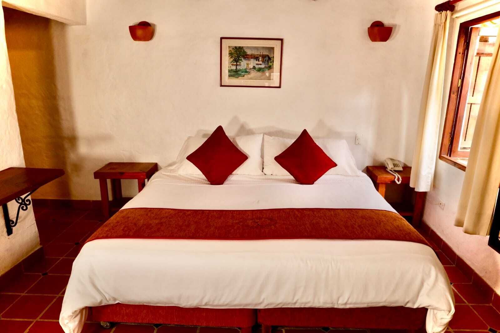 Una habitación de hotel con una cama grande, dos mesitas de noche, una lámpara de noche, un cuadro en la pared y una ventana con cortinas.