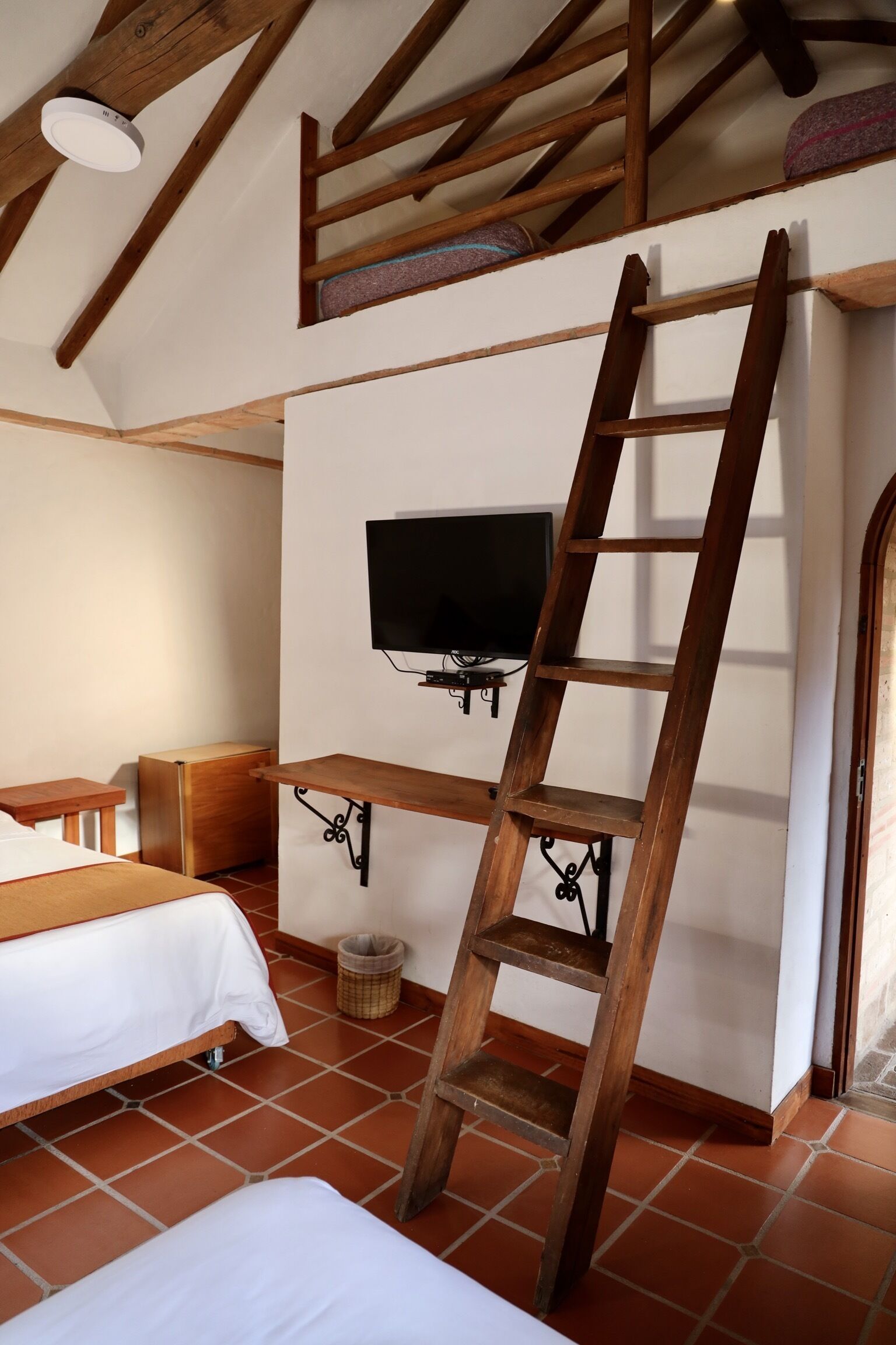 A room with a wooden ladder, a bed, a television mounted on a wall, and a small wooden cabinet.