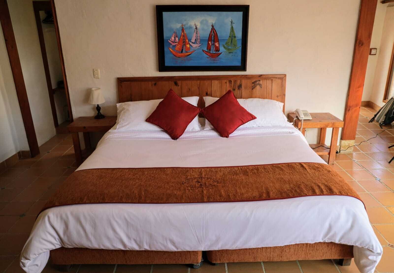 Una cama grande con cabecera de madera, dos almohadas rojas, una manta blanca y un cuadro con barcos de vela en la pared.