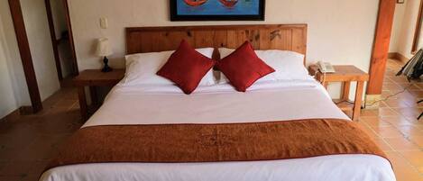 Una cama grande con cabecera de madera, dos almohadas rojas, una manta blanca y un cuadro con barcos de vela en la pared.