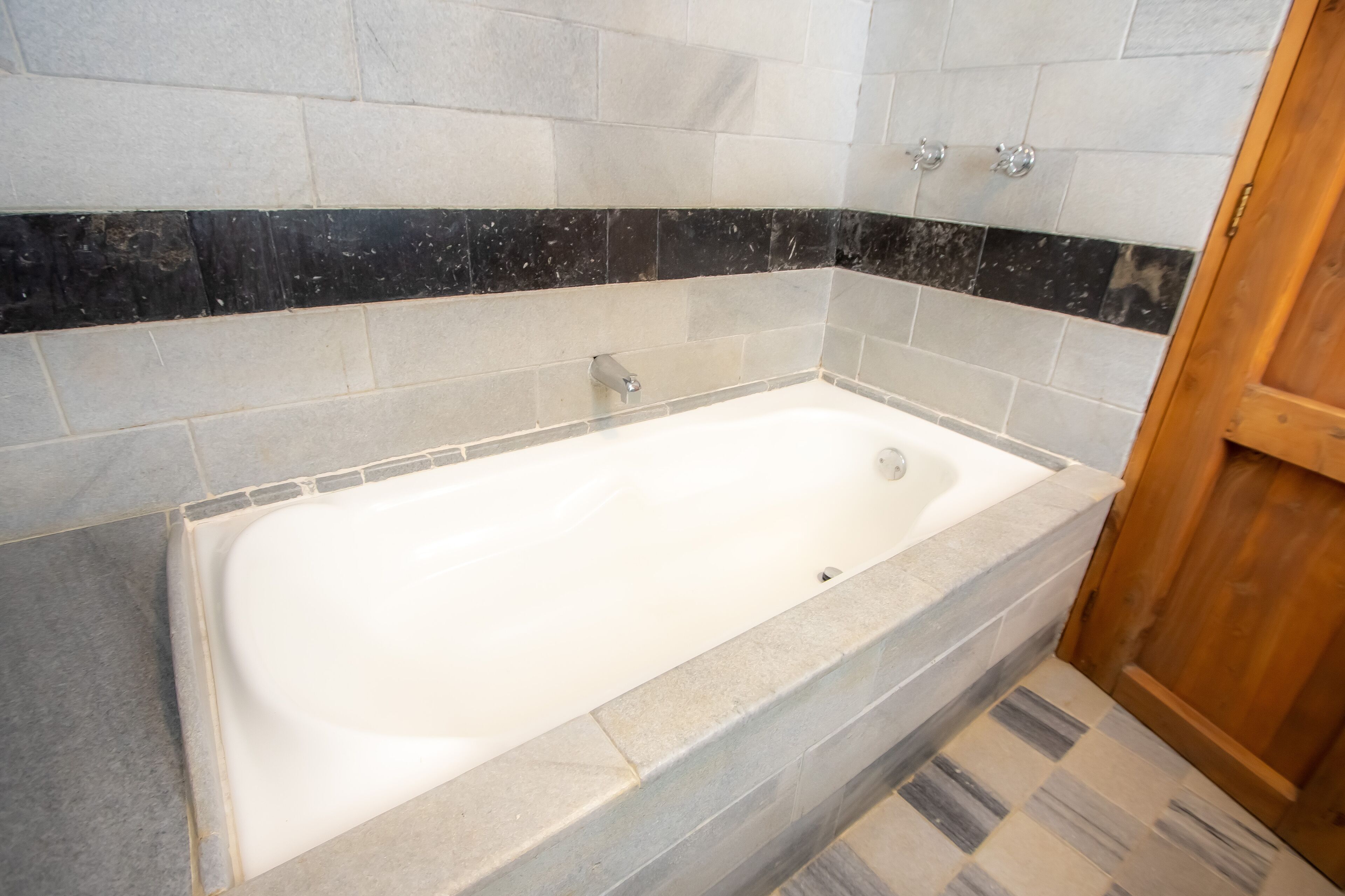 A bathroom with a white bathtub, grey and black tiles, and a wooden door.