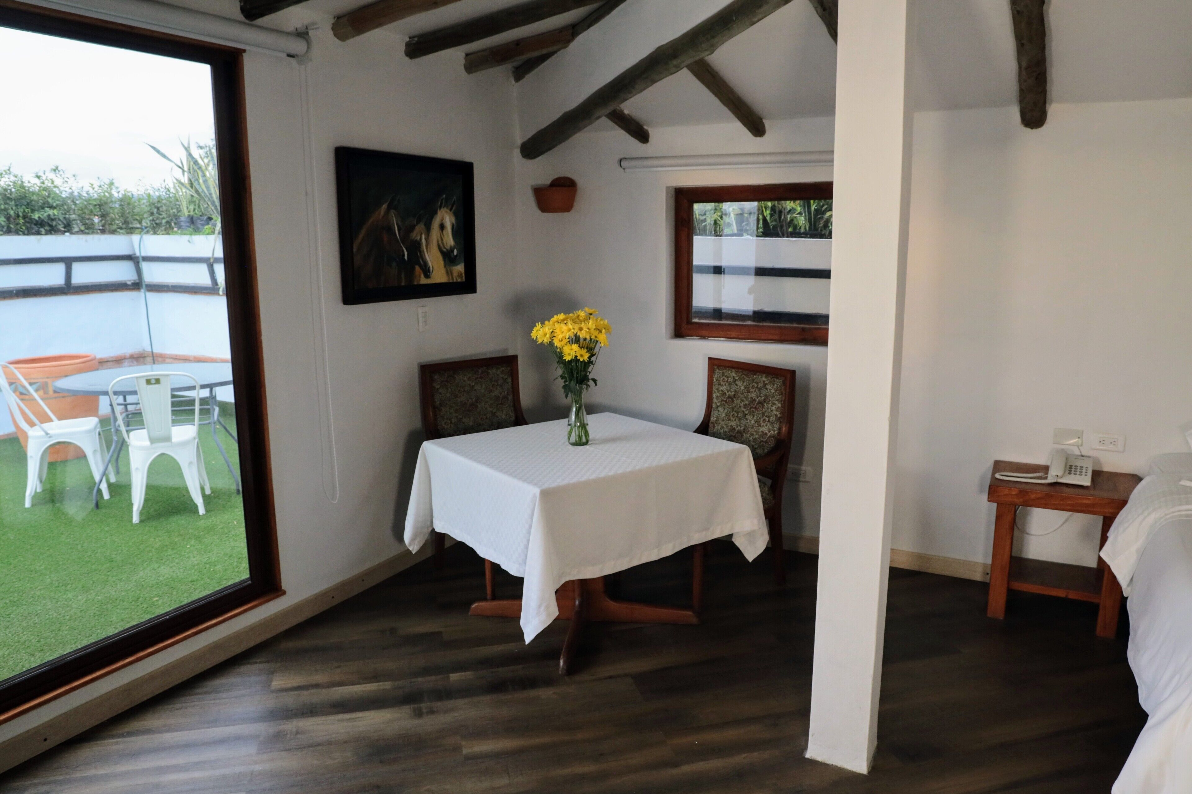 Una habitación con una mesa de comedor, sillas, un jarrón con flores amarillas, un cuadro y vistas al exterior.