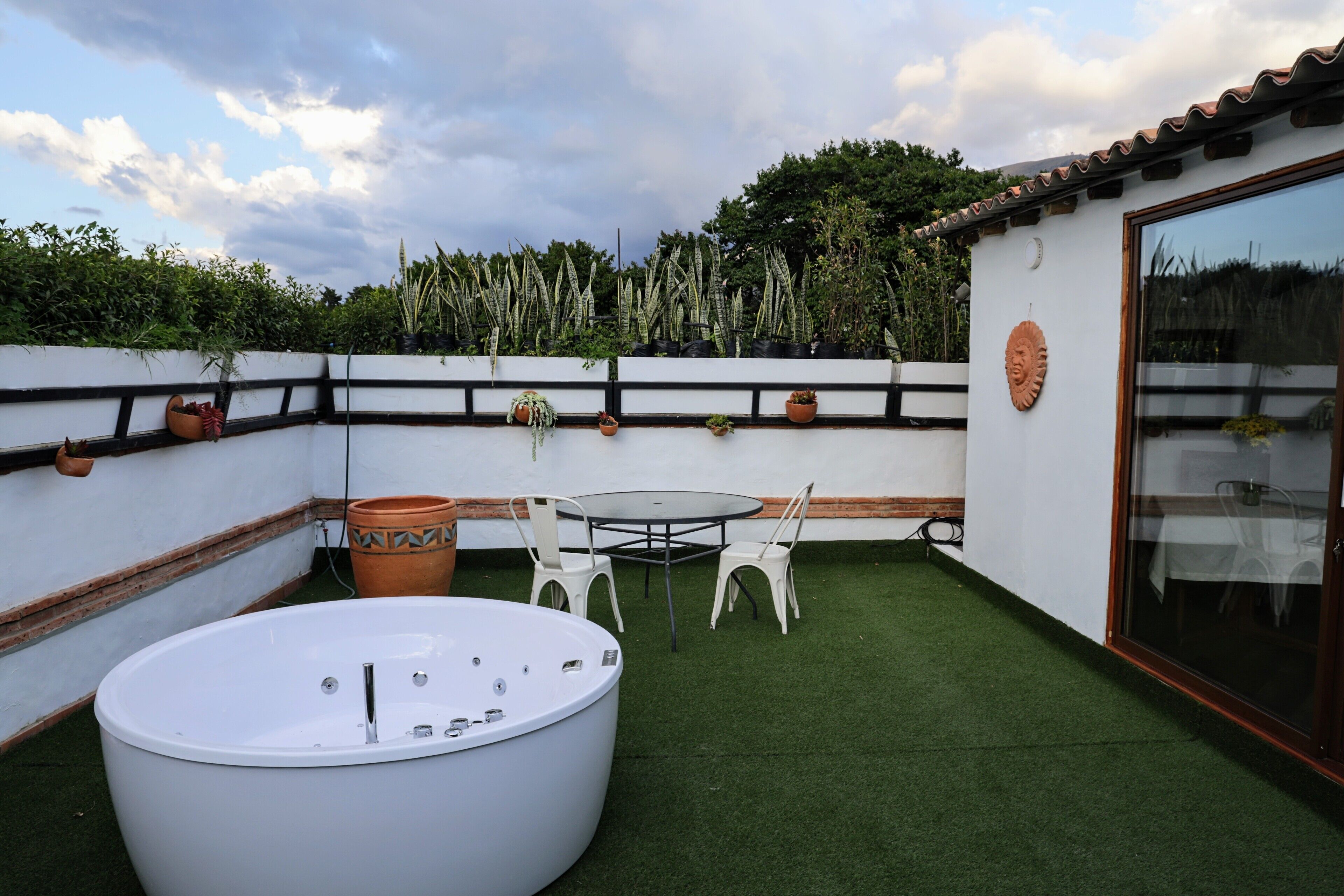 Una terraza en la azotea con un jacuzzi, una mesa redonda y sillas, rodeada de plantas en macetas y una pared blanca con detalles negros.