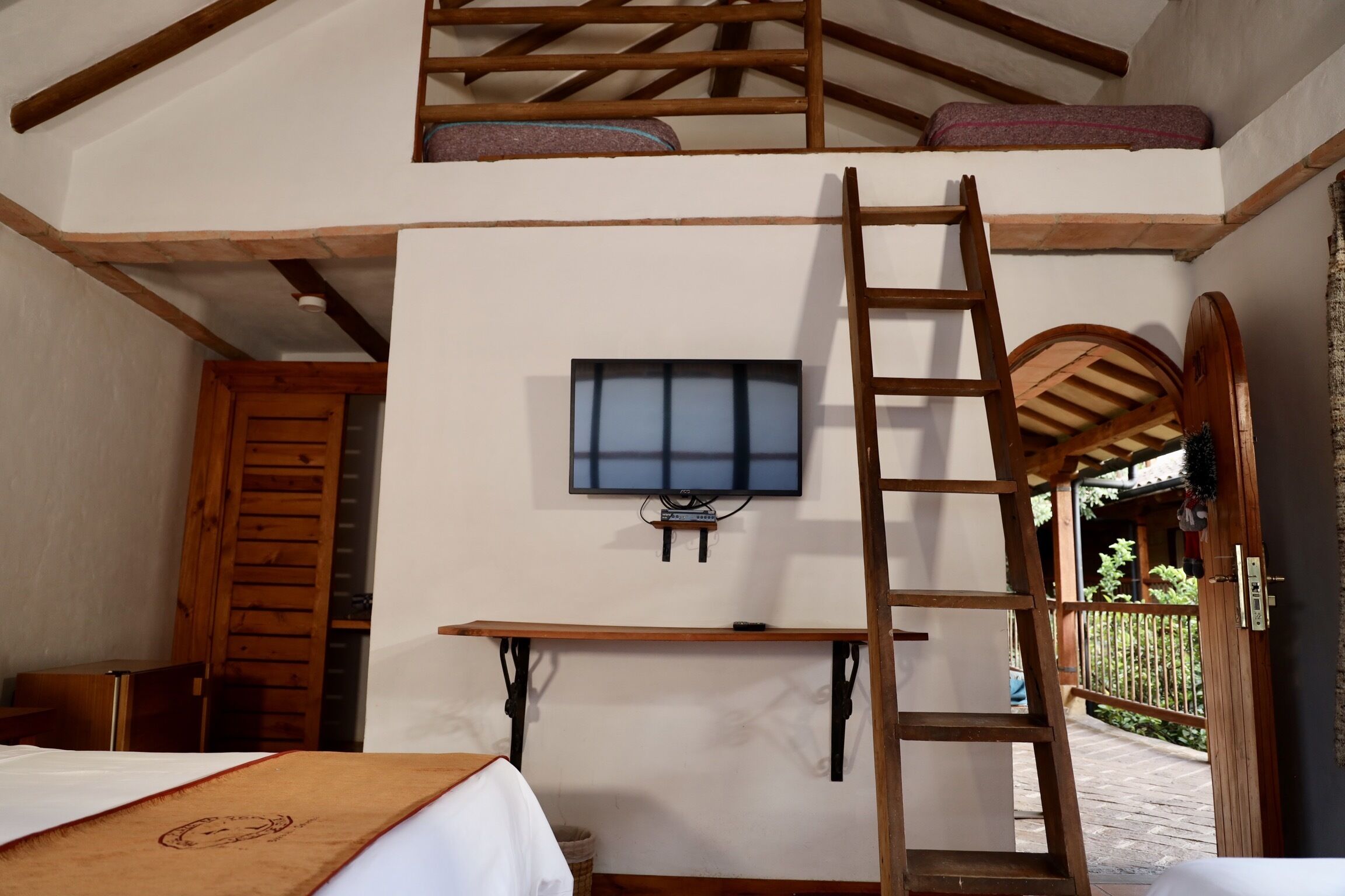 A room with a loft bed, a wooden ladder, a television mounted on the wall, and a small table.