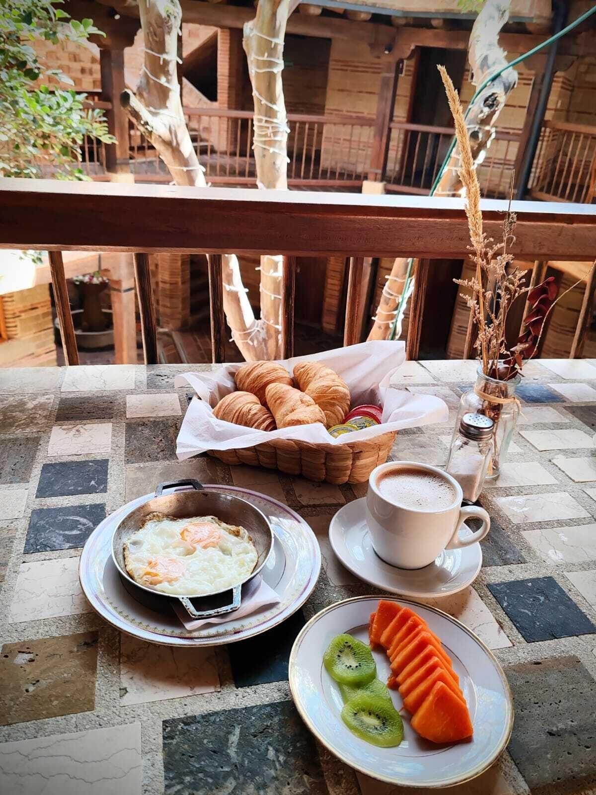 Desayuno con cruasanes, huevos, café y fruta en una terraza con barandas de madera y vista a un edificio.