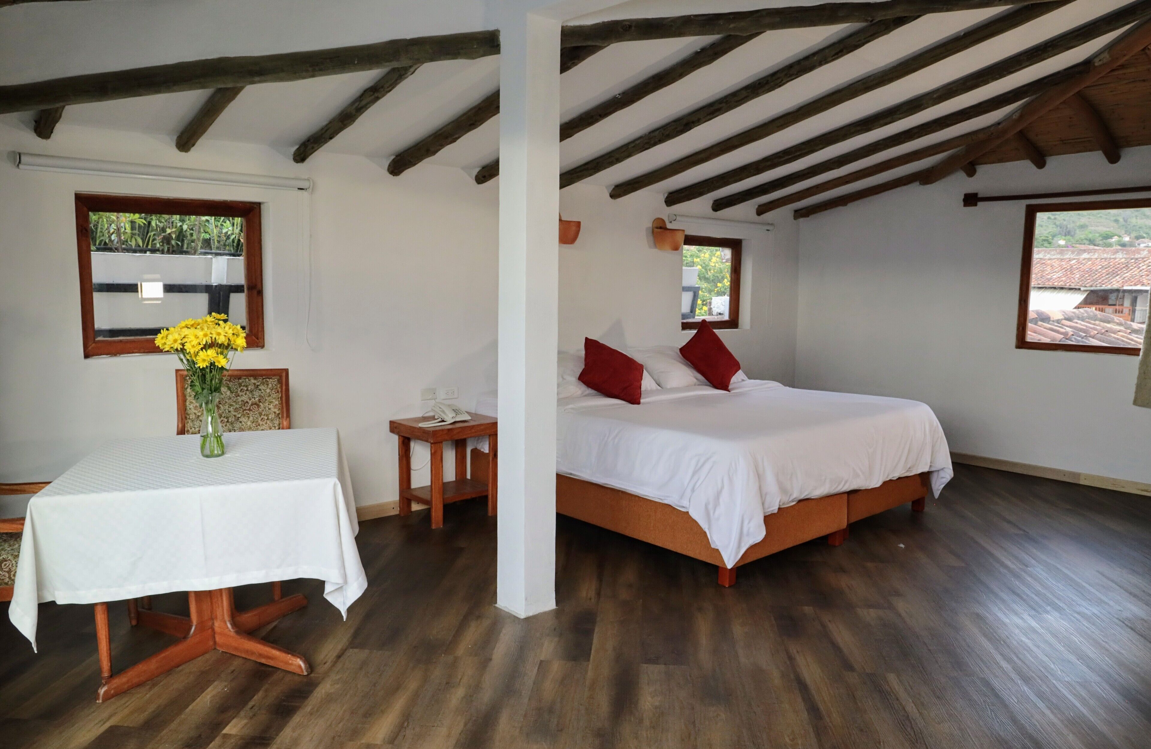 A bedroom with a wooden bed, white bedding, a small table with a vase of flowers, and wooden flooring.