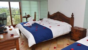 A hotel room with a bed, a wooden nightstand, a desk with a chair, and a window with a view of greenery. - Hotel Campestre Bachue (Villa de Leyva)